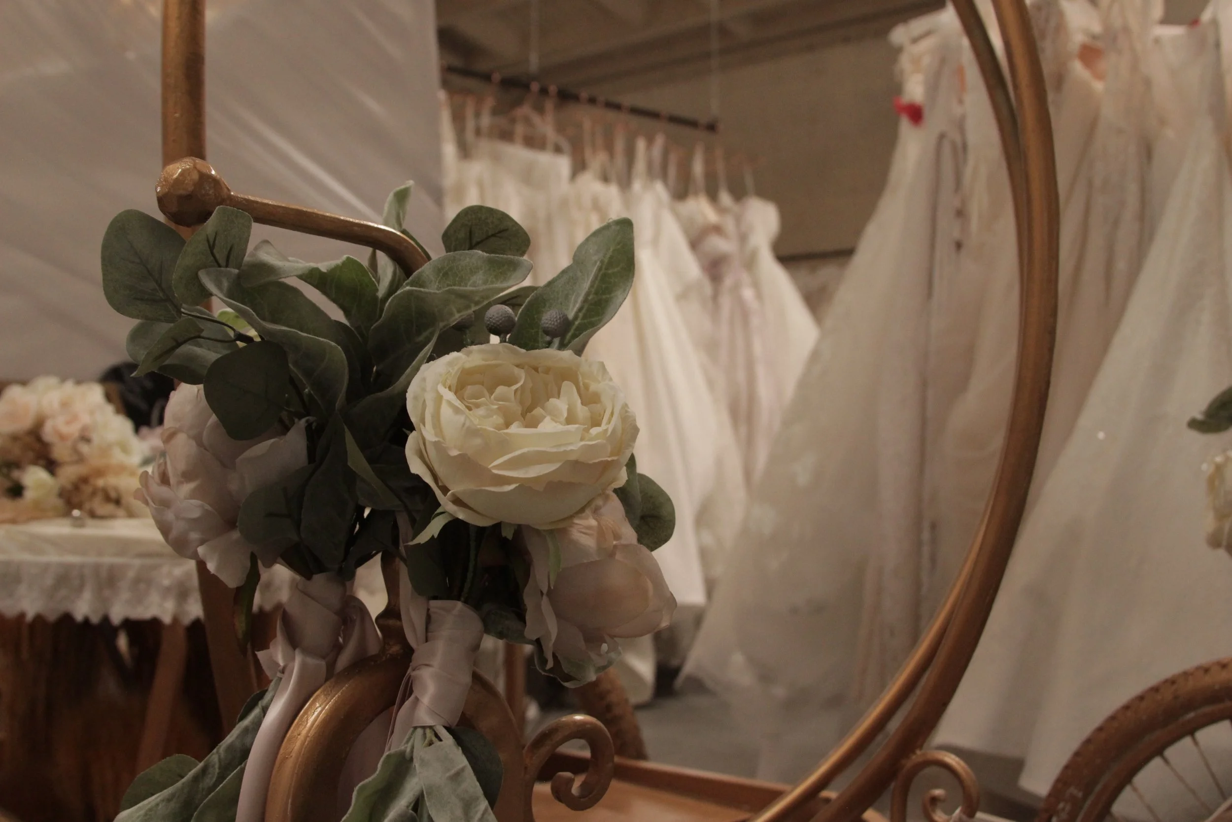 Close-up of a floral arrangement with white roses and green leaves, reflected in a rounded mirror hanging on a wall with wedding dresses in the background.