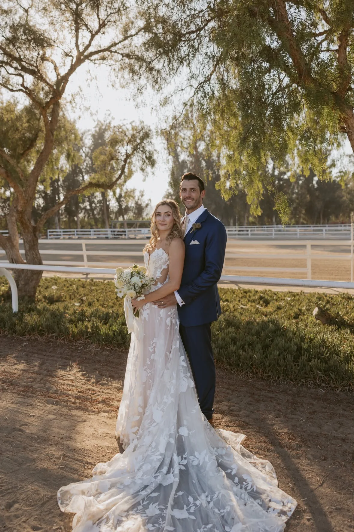 A bride and groom standing together outdoors, with the bride holding a bouquet of white flowers. They are dressed in wedding attire, smiling and posing for a photo in a scenic setting with trees and a fence in the background during sunset.