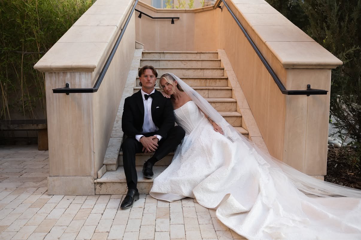 A bride and groom sitting on stairs outdoors, with the bride resting her head on the groom's shoulder. The groom wears a black tuxedo and bow tie, and the bride wears a white wedding gown with a long train and veil.