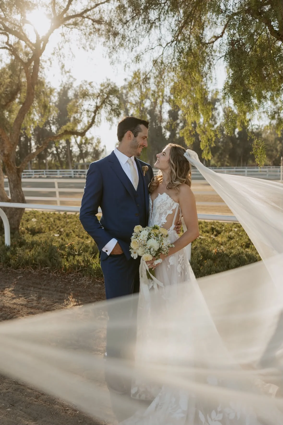 A bride and groom standing outdoors near trees, smiling and looking at each other, with sunlight filtering through the leaves. The bride is holding a bouquet and wearing a wedding dress with a flowing veil, while the groom is in a blue suit.