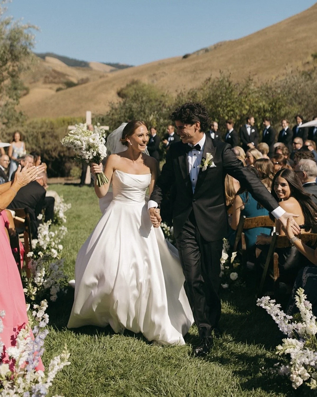 A bride and groom holding hands and smiling during an outdoor wedding ceremony, surrounded by seated guests and scenic hills in the background.