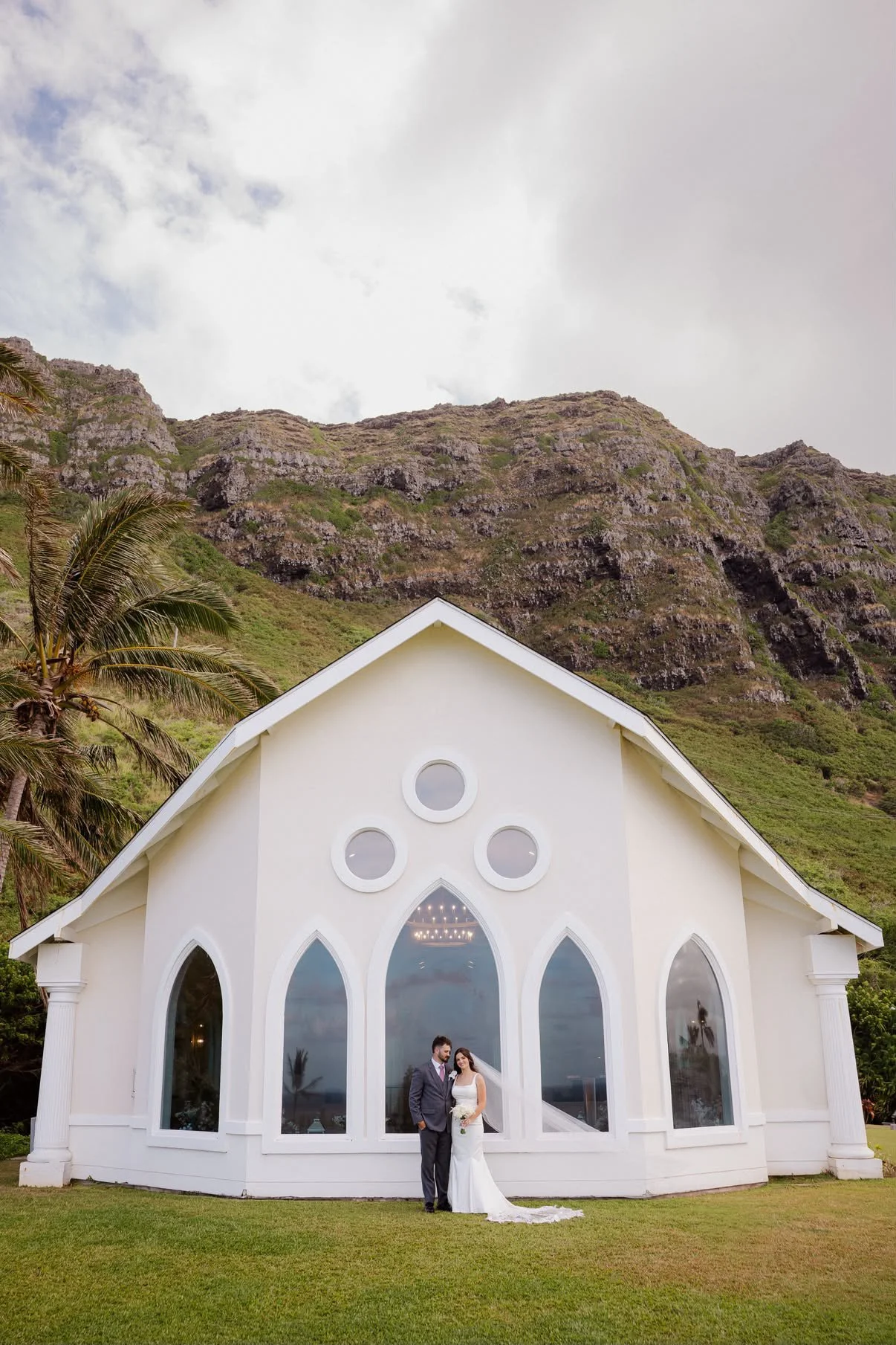 A bride and groom standing outside a white church with large arched windows, lush green grass in front, tall palm trees and a mountain with rocky slopes and green vegetation in the background.