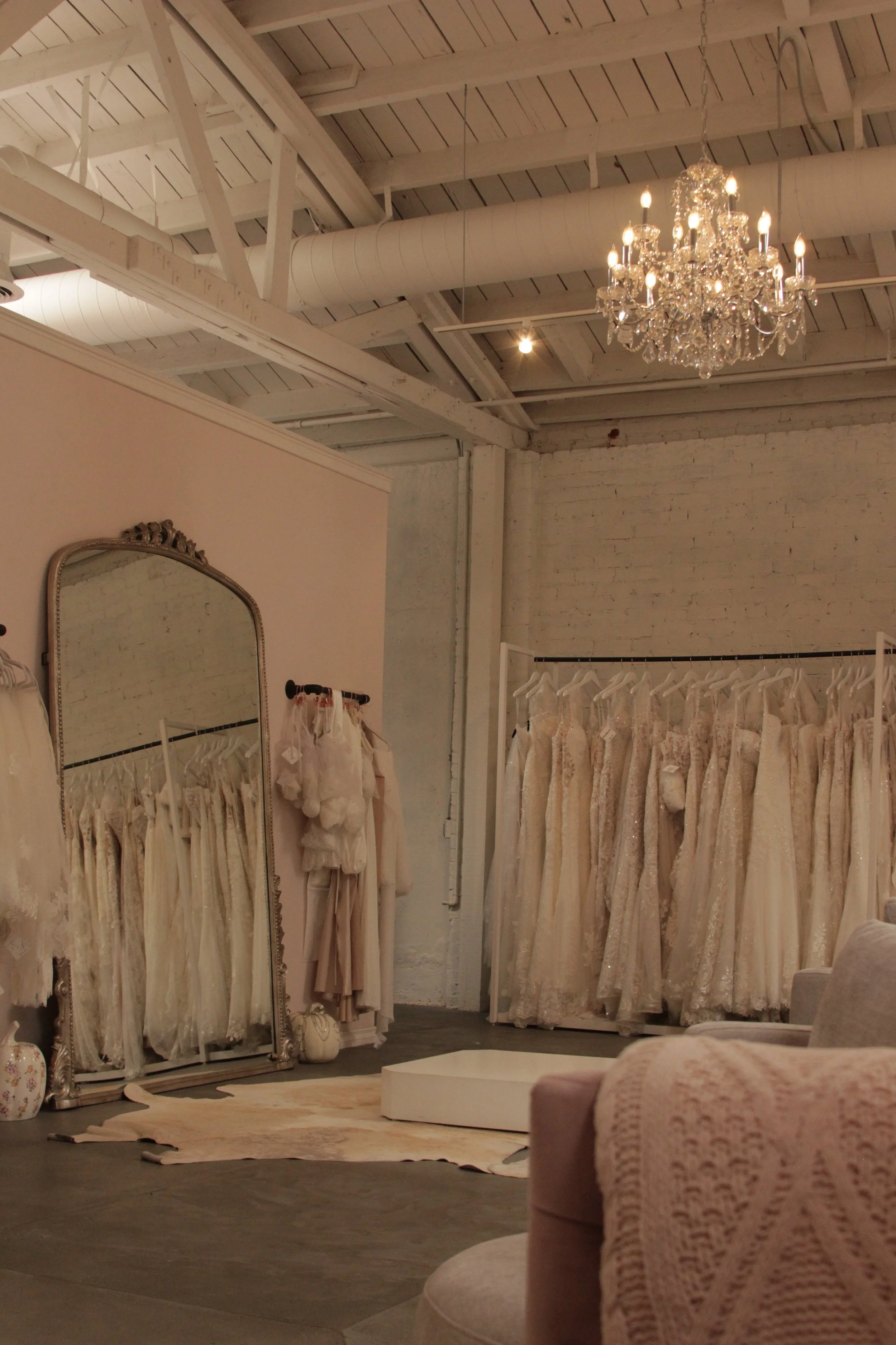 Interior of a bridal boutique featuring a display of wedding dresses on racks, a large ornate mirror, a chandelier hanging from the ceiling, and a cozy seating area with pillows.
