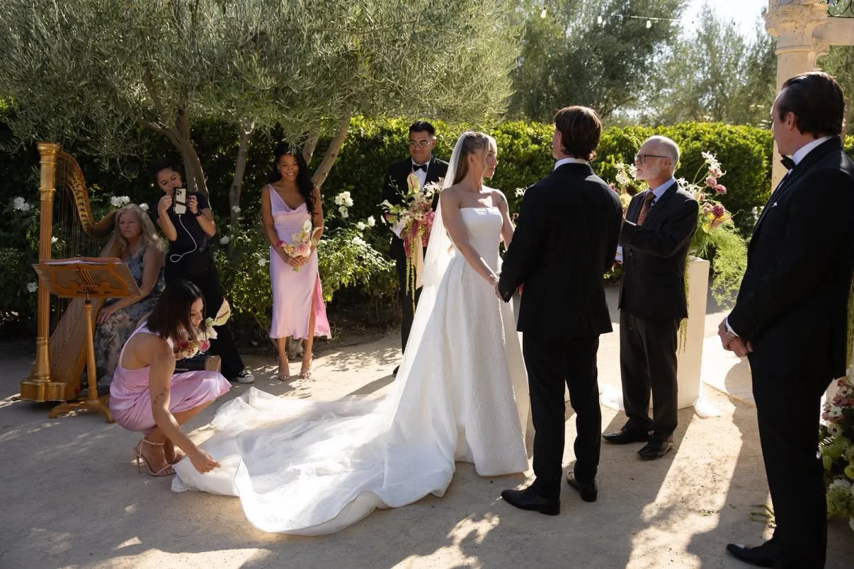 A wedding ceremony outdoors with a bride and groom holding hands, surrounded by bridesmaids, groomsmen, family, and friends, with a harp and floral decorations in the background.