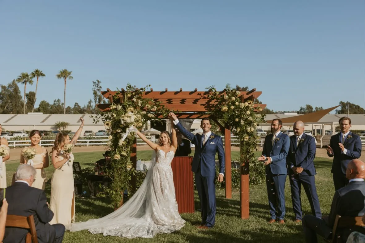A wedding ceremony outdoors with a bride and groom holding hands under a floral arch, surrounded by bridesmaids and groomsmen, with guests seated nearby.