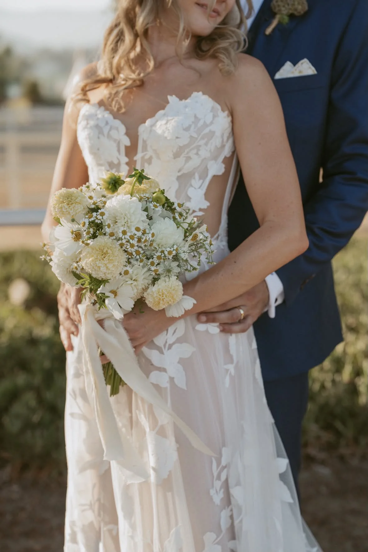 A bride and groom embracing outdoors, the bride holding a bouquet of white flowers and wearing a white lace wedding gown, the groom in a dark suit. They are close together on a sunny day.