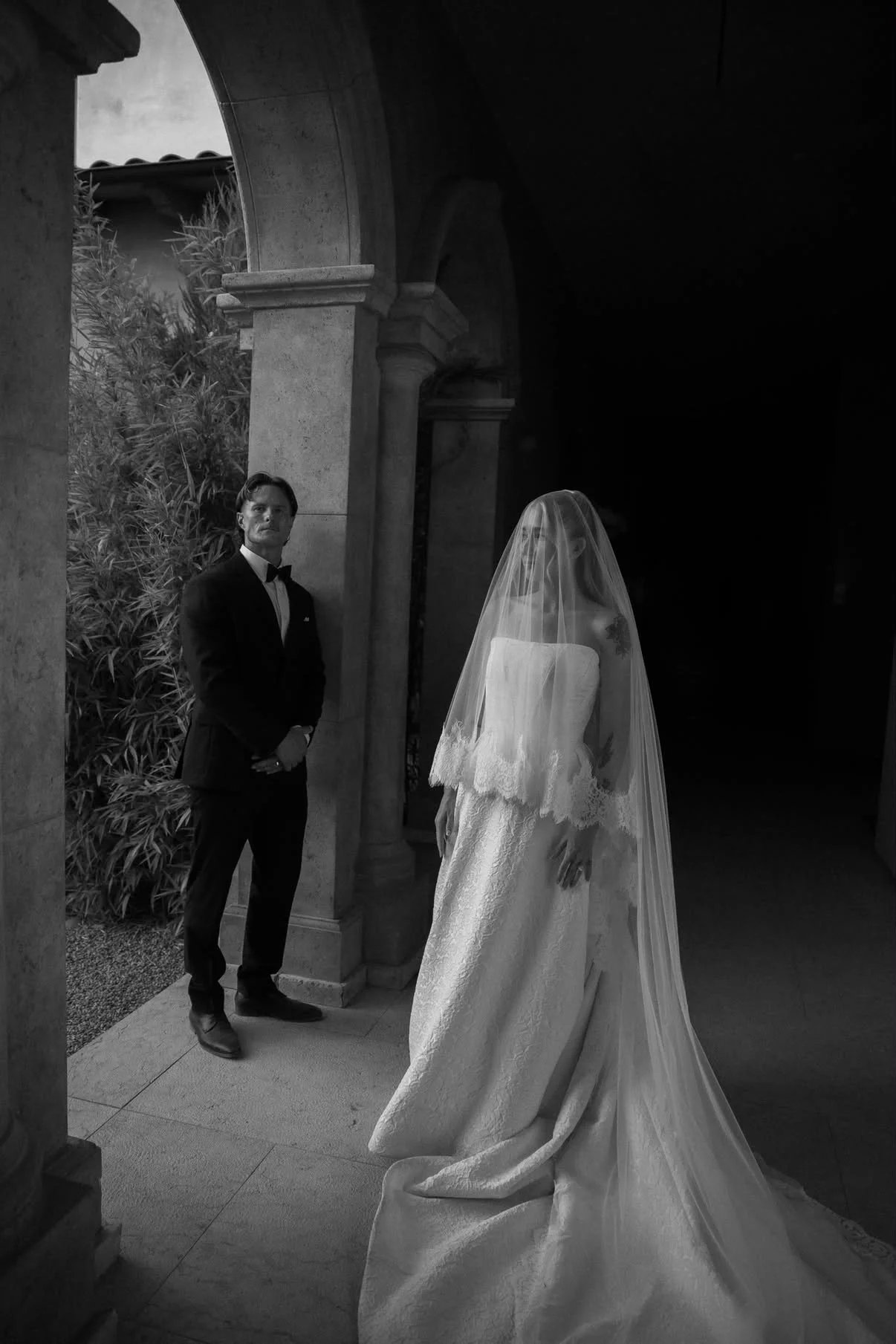 Black and white photo of a bride and groom standing outside a building with columns. The groom is in a tuxedo, looking at the bride, who is wearing a wedding dress and veil.