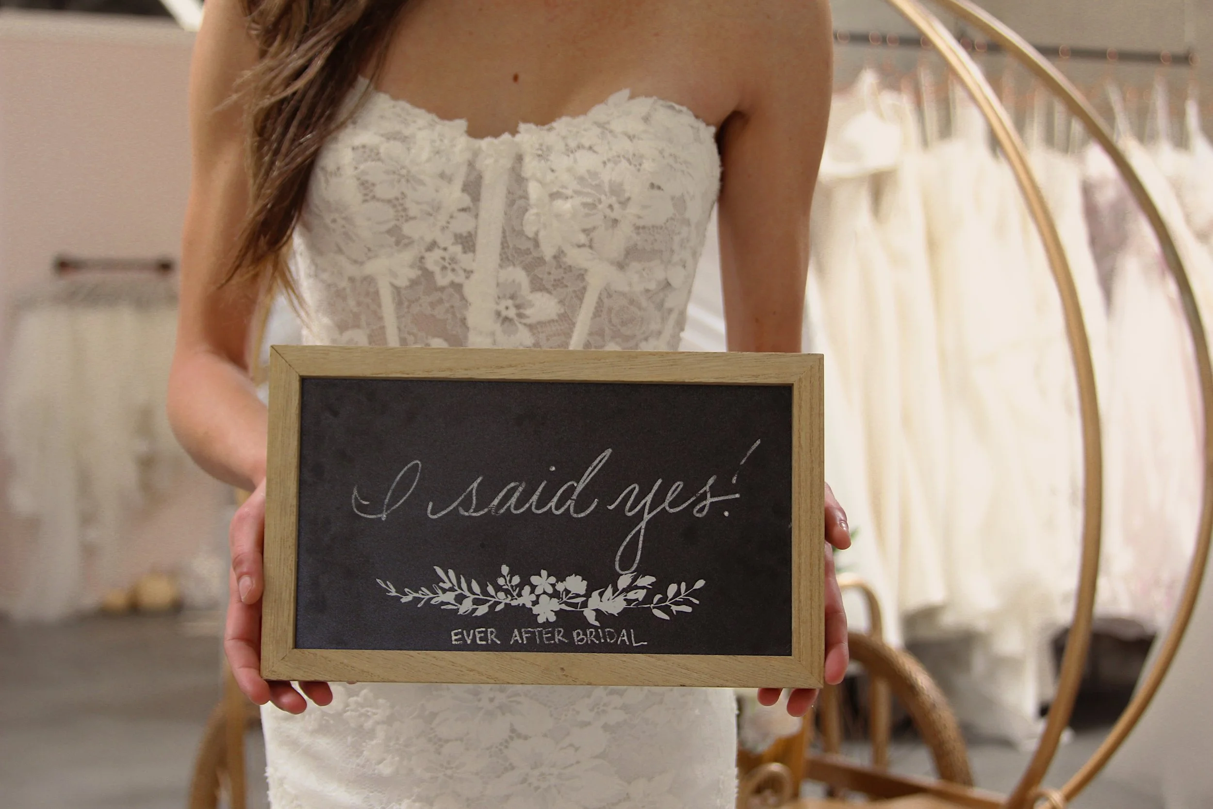 Person in a wedding dress holding a chalkboard sign that says 'I said yes' with floral decoration and 'Ever After Bridal' written at the bottom.
