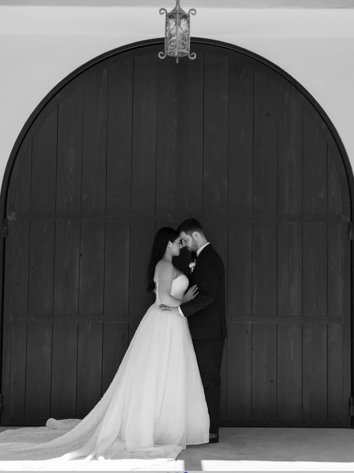 A black and white photo of a bride and groom standing close with foreheads touching, in front of a large wooden arched door, with a decorative lantern hanging above.