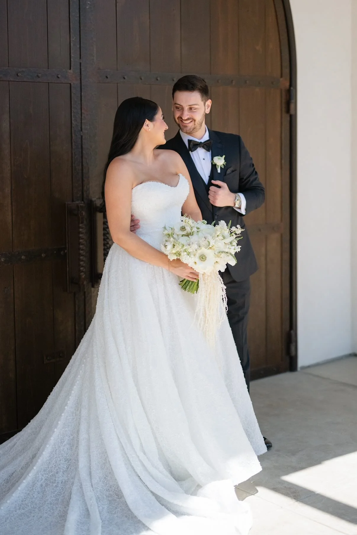 A bride and groom smiling at each other, standing in front of a wooden door. The bride is holding a bouquet of white flowers and wearing a white strapless wedding gown. The groom is dressed in a black tuxedo with a bow tie and boutonniere.