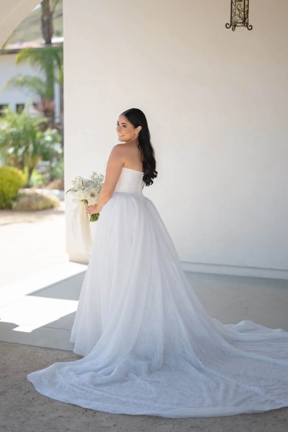 A woman in a strapless white wedding gown holding a bouquet of white flowers, smiling, with long dark hair, standing outside near a white wall and greenery.