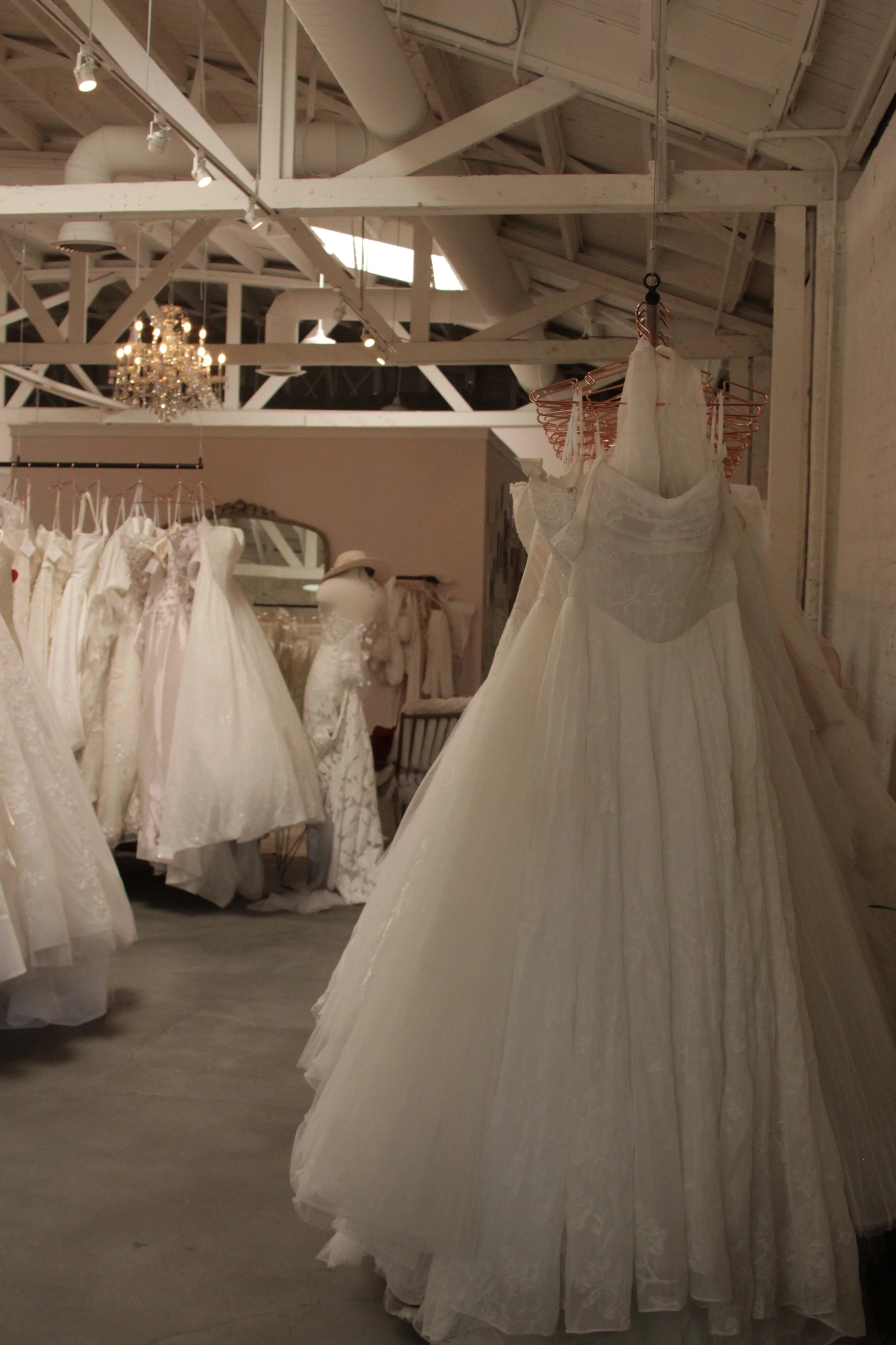 Wedding dresses hanging on racks in a bridal store, with a chandelier visible in the background and a rustic ceiling with exposed beams and ductwork.