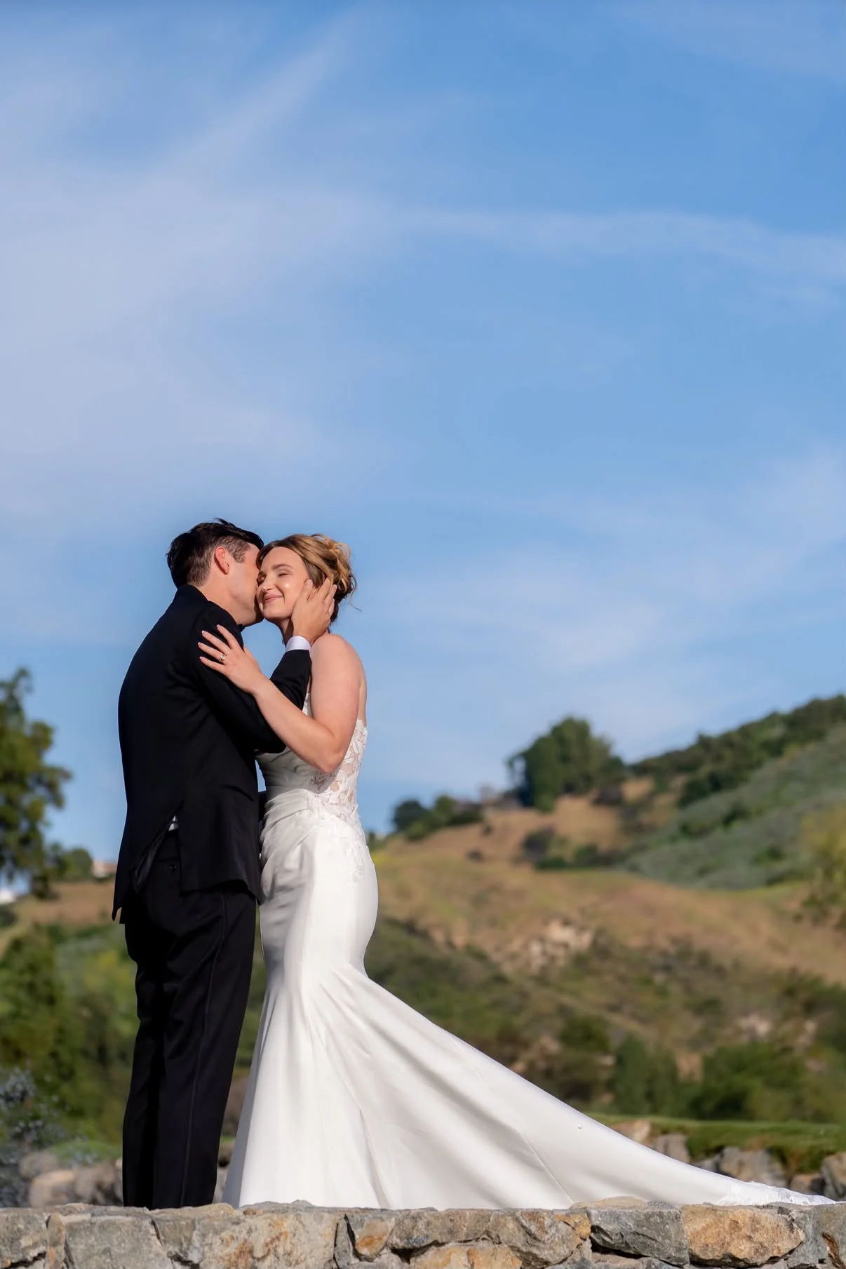 A newlywed couple sharing an intimate moment outdoors during the daytime, with rolling hills and a blue sky in the background.