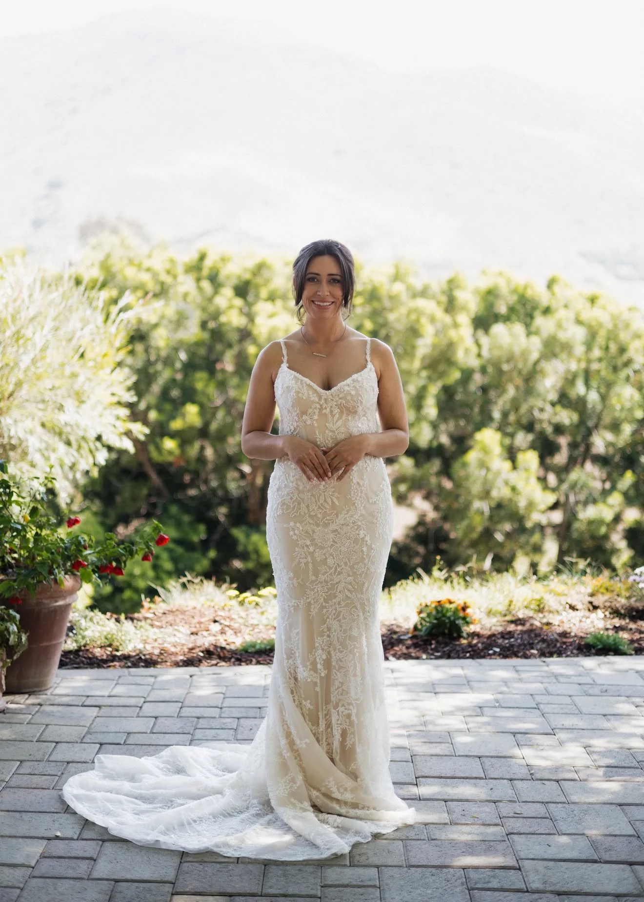 A woman in a wedding dress standing outdoors on a stone patio with greenery and flowers in the background.