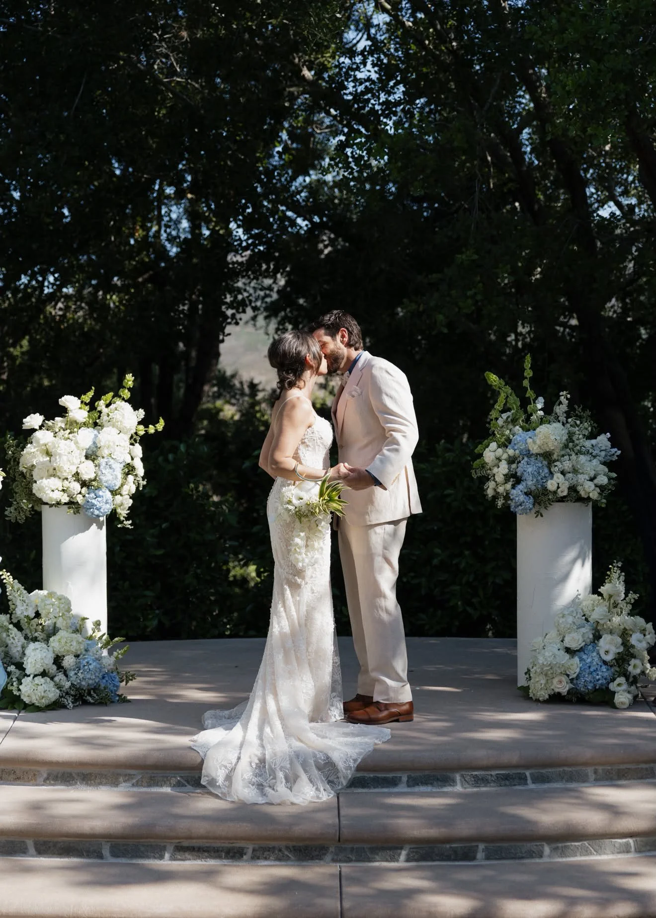 A bride and groom sharing a kiss during their outdoor wedding ceremony, surrounded by white and blue floral arrangements in an outdoor garden setting.