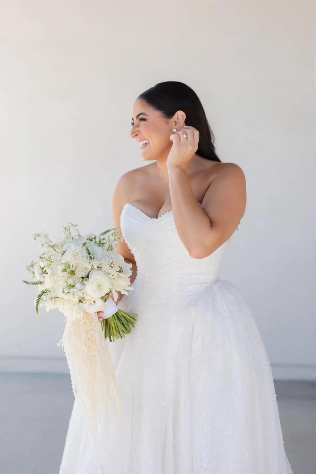 A woman wearing a strapless white wedding dress holding a bouquet of white flowers while smiling and touching her earring.