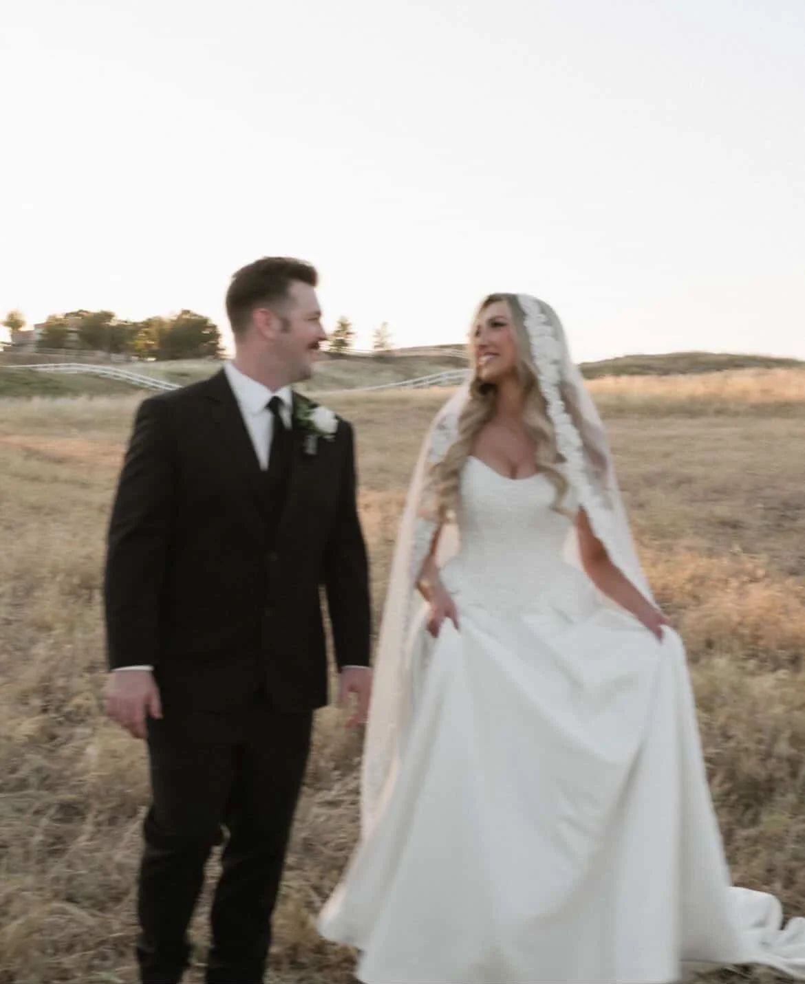 A bride and groom smiling at each other outdoors during sunset, with a grassy field and trees in the background.