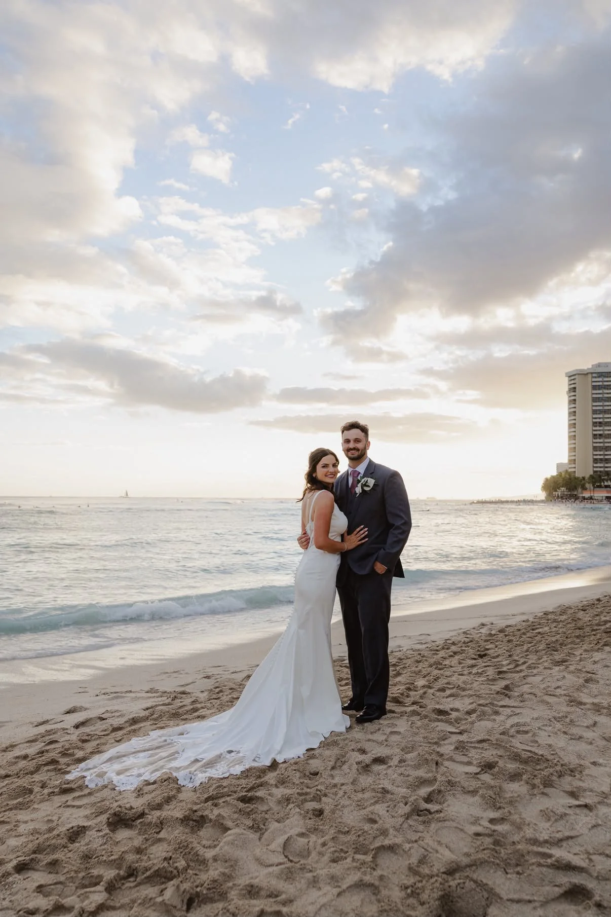 A bride and groom in wedding attire standing on a beach with the ocean and a city skyline in the background during sunset.
