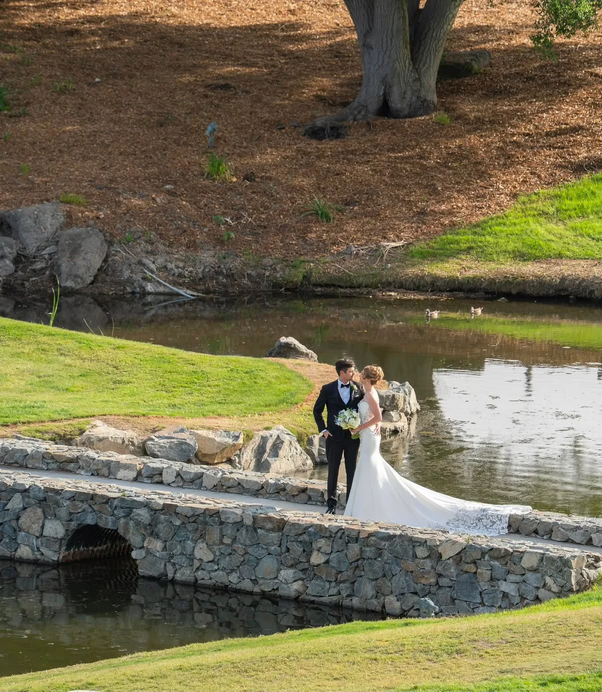 A bride and groom in wedding attire standing on a stone bridge by a pond, kissing with ducks swimming in the background.