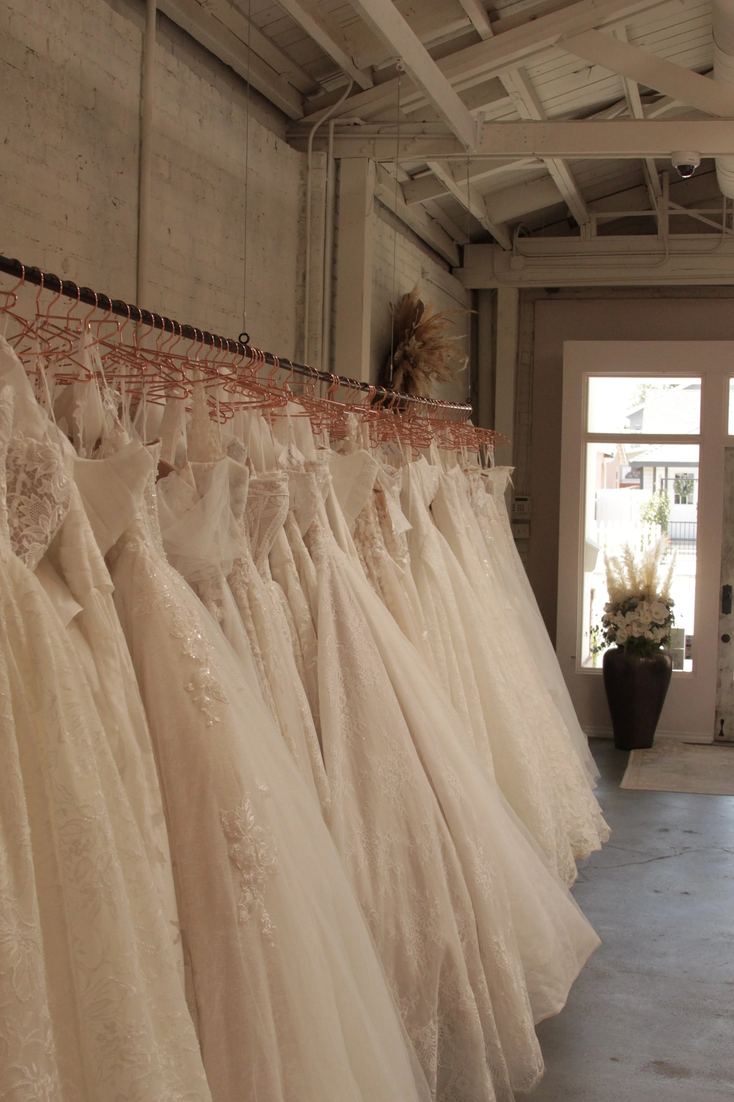 A row of white wedding dresses on pink hangers inside a boutique with a brick wall and a large window.