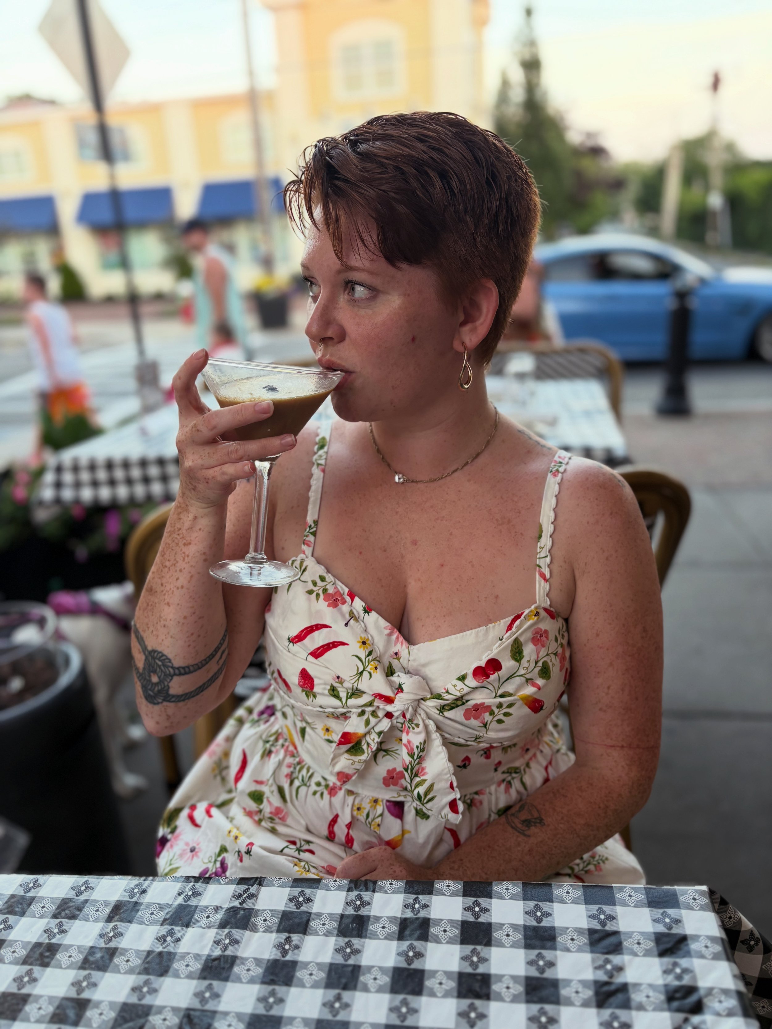 A woman with short reddish-brown hair and freckles, wearing a floral dress with spaghetti straps, is sitting at a checkered table outdoors, holding a cocktail glass near her mouth. In the background, there are blurred pedestrians, a light yellow building with blue awnings, and a parked blue vehicle.