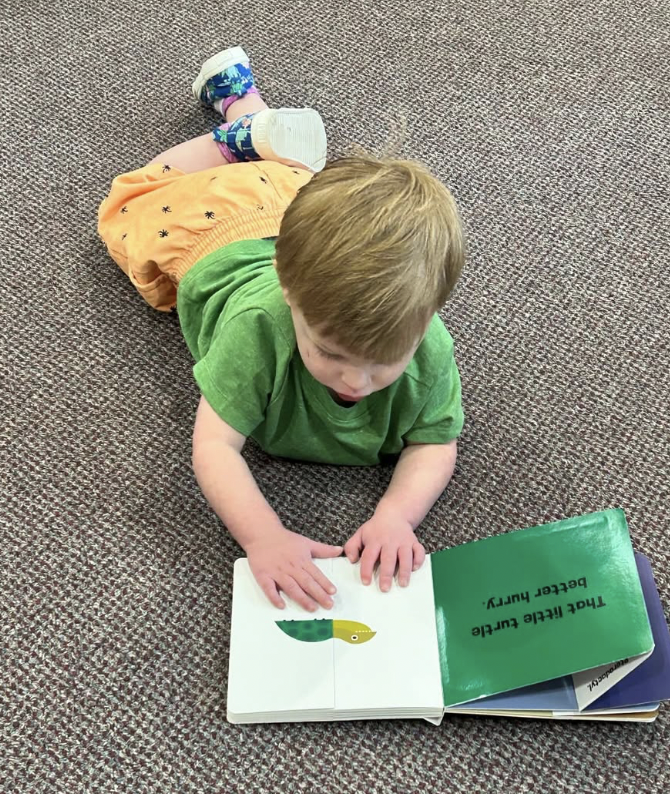 A young boy lying on a carpeted floor looking at a book about a tickle bug.