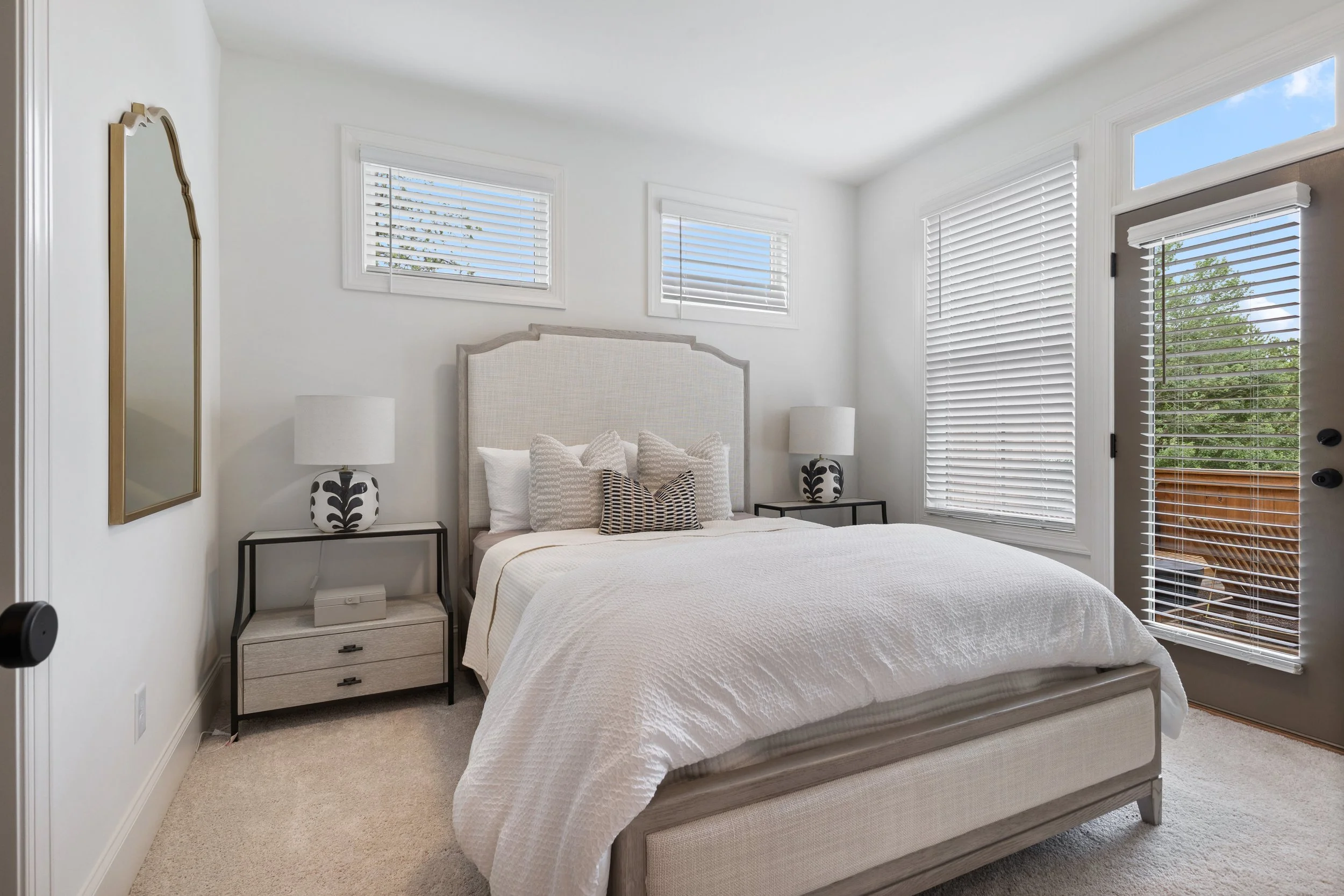 Bright bedroom with white walls, a plush bed with a textured white bedspread, and decorative pillows. Two black nightstands with white lamps featuring black patterns flank the bed. There are three windows with white blinds allowing natural light, and a door with blinds leading to an outdoor balcony. A tall mirror with a gold frame leans against the wall.