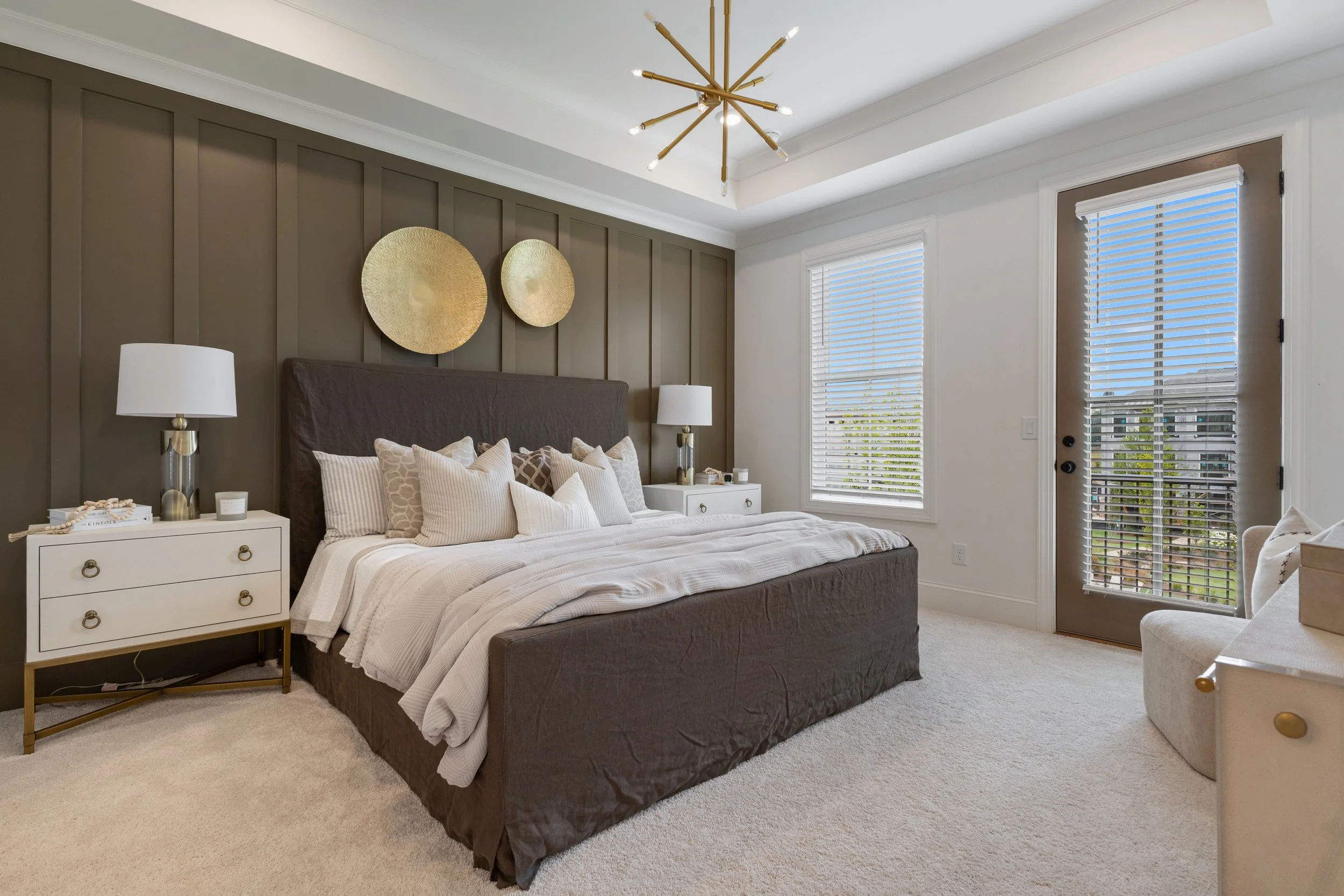 A modern bedroom with a dark upholstered bed, white bedding, and multiple decorative pillows. Two white nightstands with gold accents and table lamps are on either side of the bed. The wall behind the bed has vertical paneling and two large round gold wall decor pieces. There are two windows with white blinds and a door with white blinds leading outside. The ceiling features a gold modern chandelier with multiple arms.
