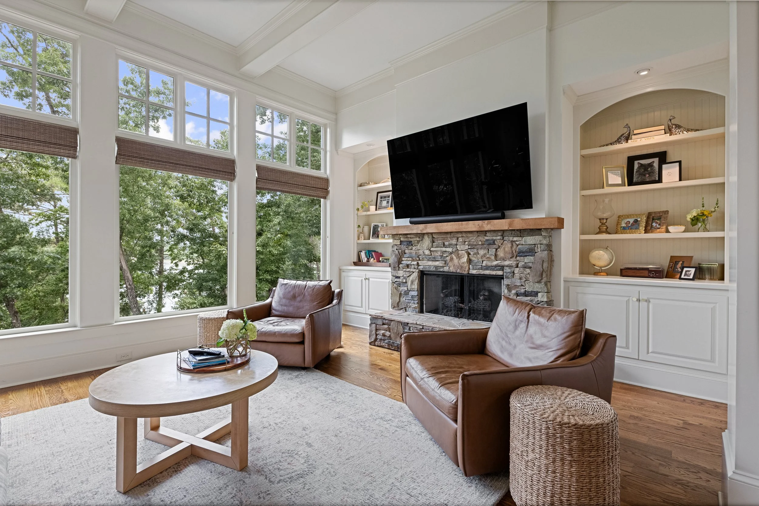 Living room with large windows, a stone fireplace, a wall-mounted TV, white built-in shelves with decorative items, and leather chairs.