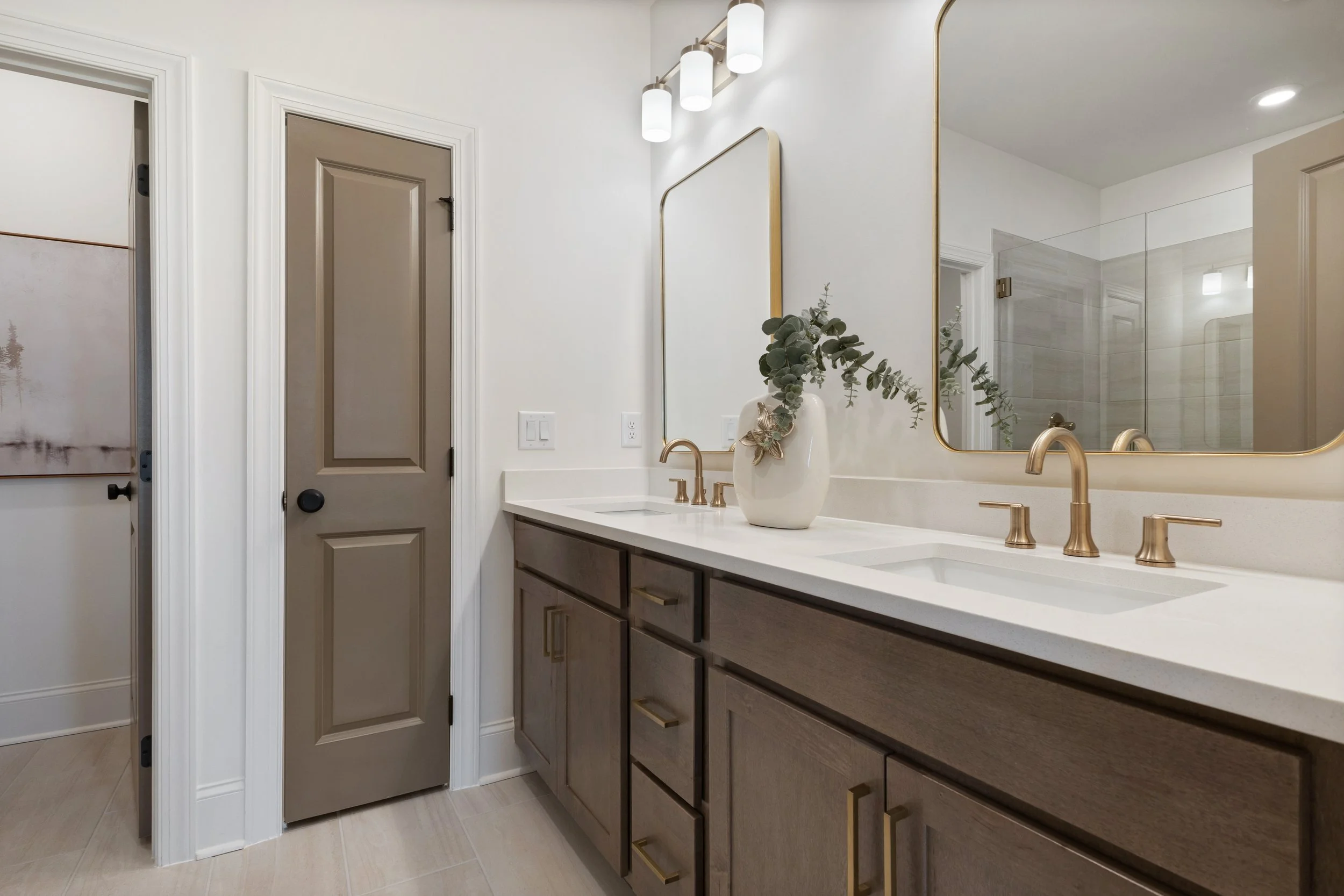 Bathroom with double sinks, large mirrors with gold trims, a vase with greenery on the counter, white walls, a door, and a shower with glass door.