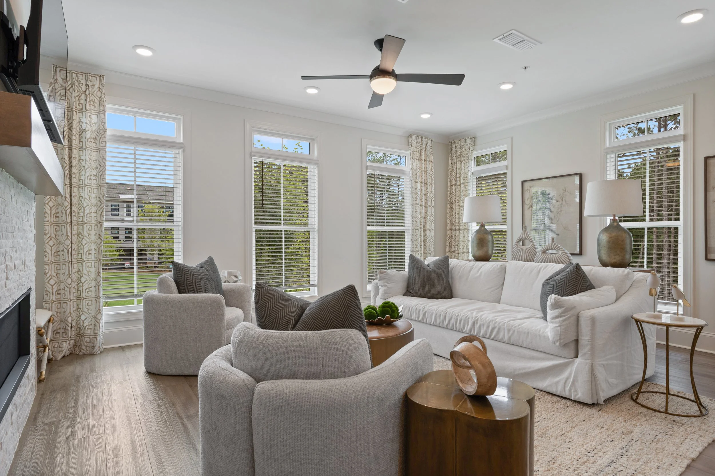 Bright modern living room with large windows, white walls, light wood flooring, a white sofa, upholstered armchair, decorative lamps, and ceiling fan.