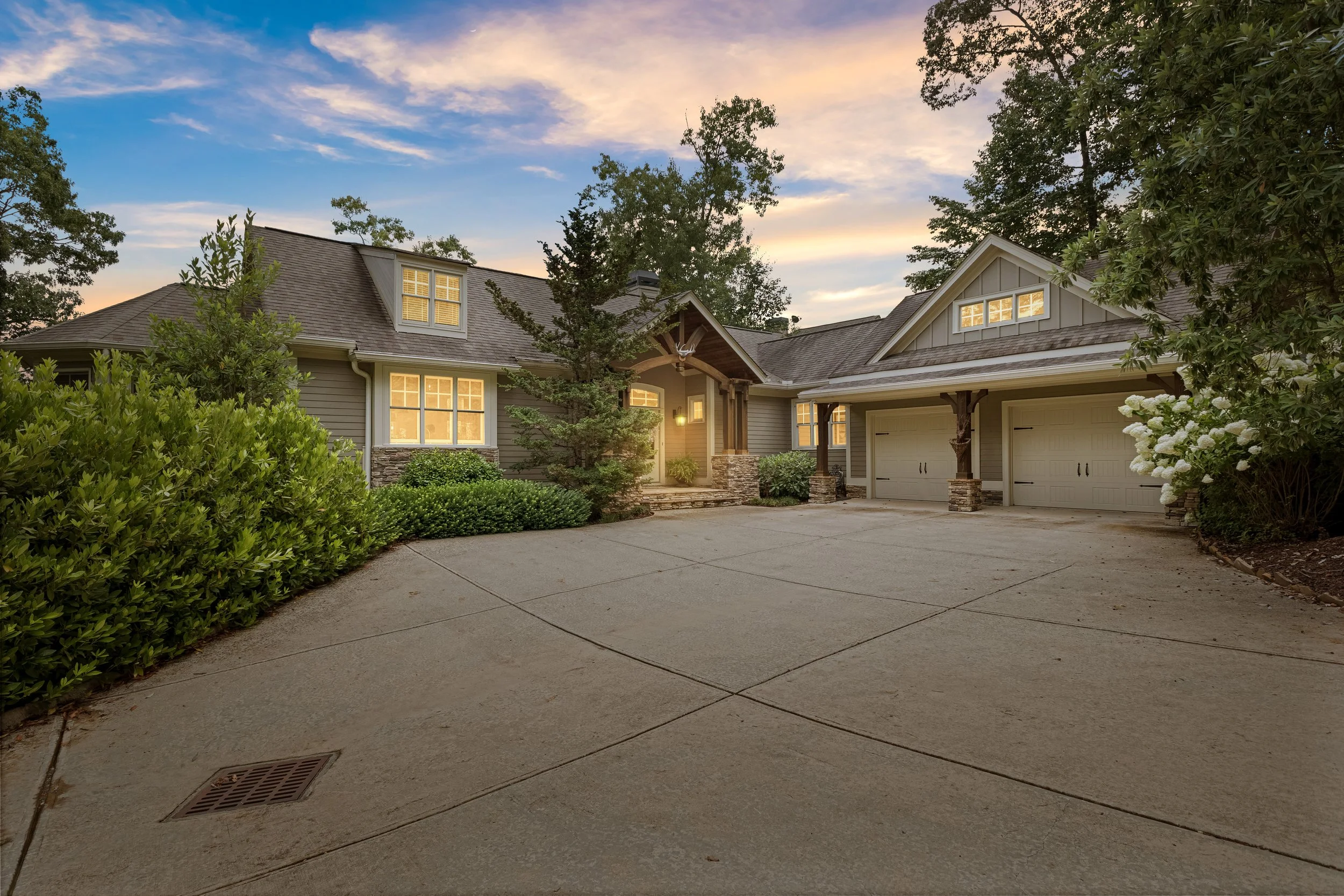 View of a suburban house during sunset with lit windows, a driveway, trees, bushes, and a partly cloudy sky.