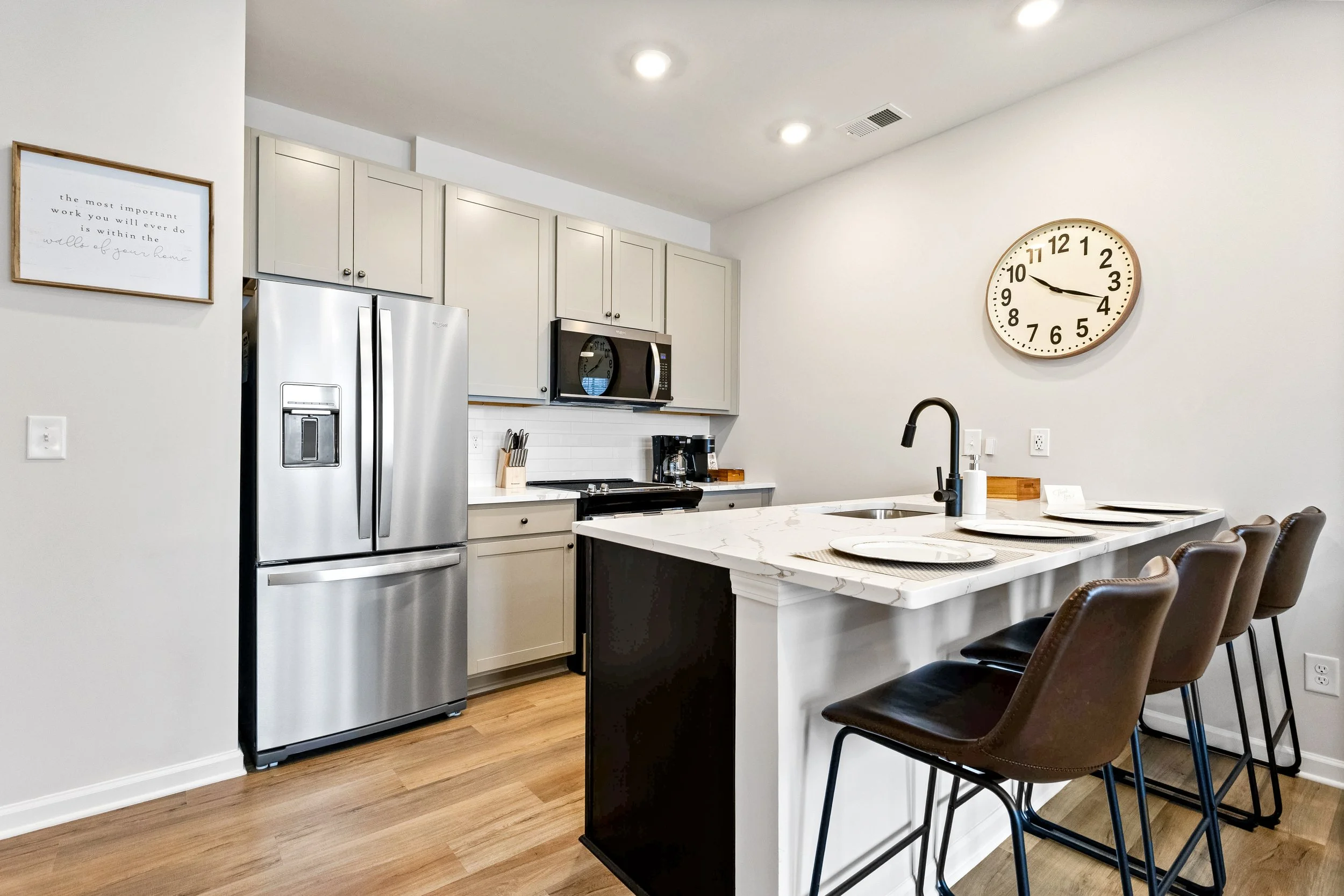 Modern kitchen with stainless steel fridge, microwave, white cabinets, black countertop, kitchen island with white marble top, hanging chairs, wall clock, wall art, and hardwood flooring.