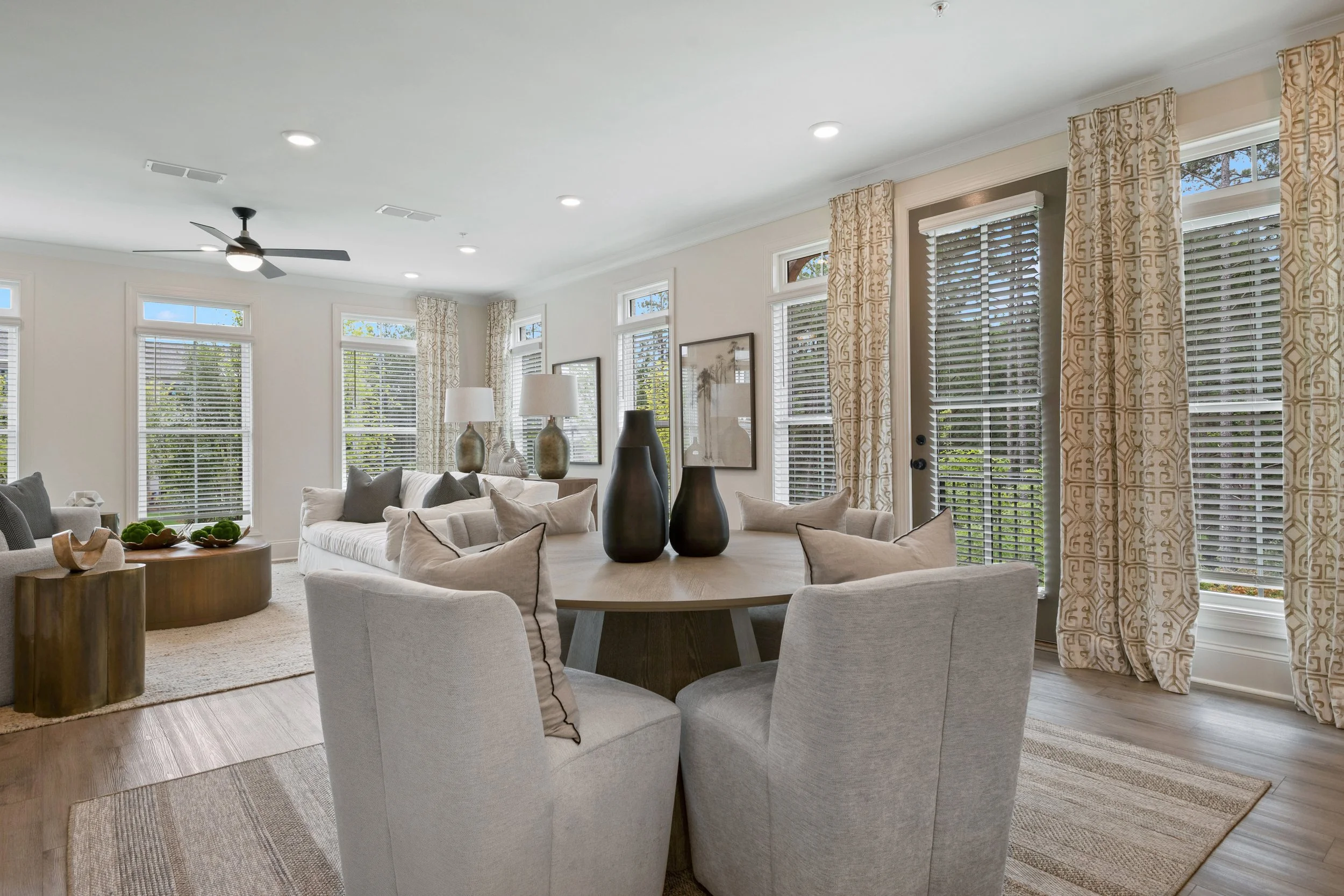 Living room with large windows, beige curtains, gray sofa, round wooden dining table with beige chairs, decorative vases, hanging wall art, ceiling fan, and recessed lighting.