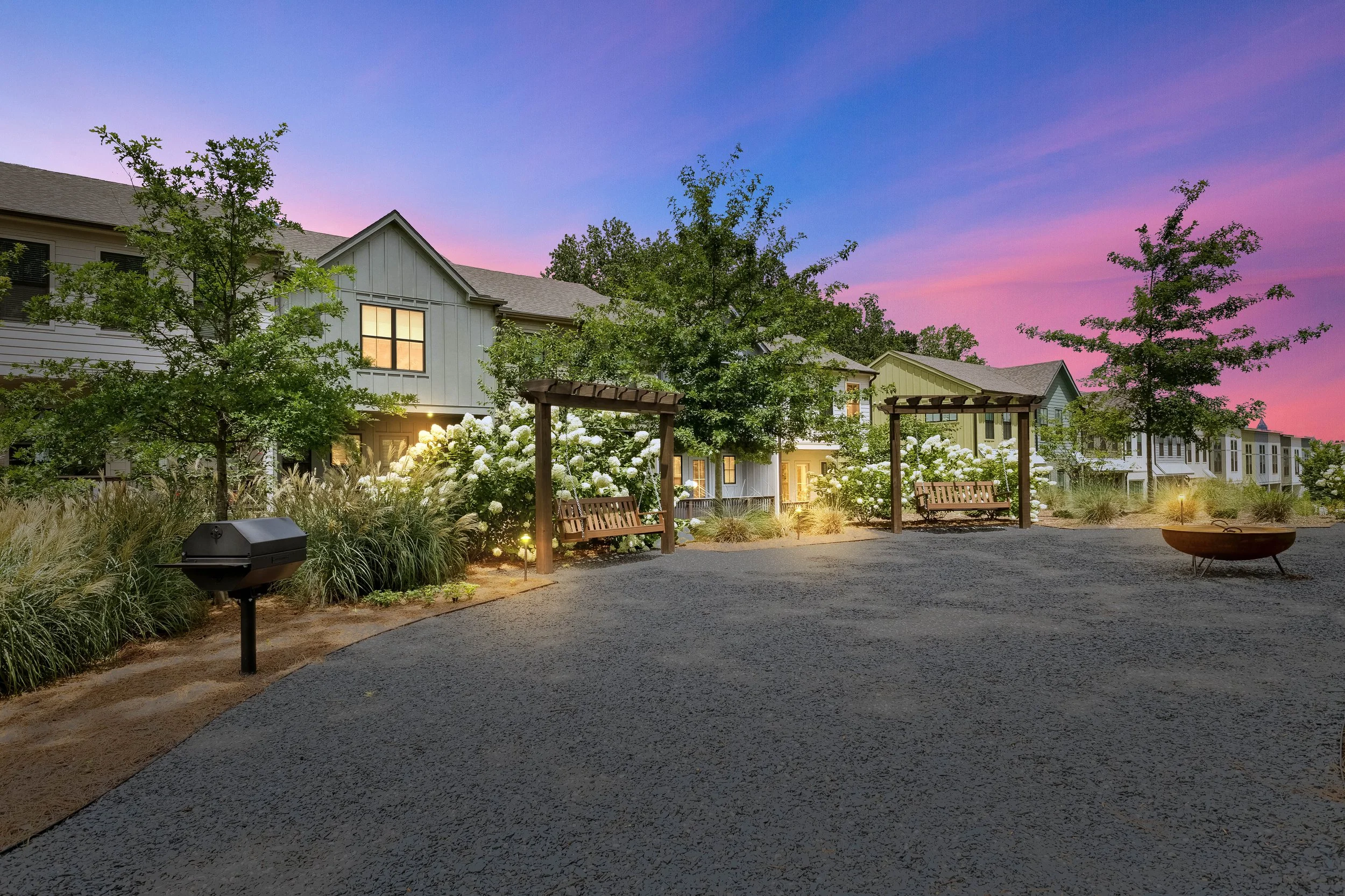 Residential complex with modern houses, trees, benches, gravel pathways, a barbecue grill, and sunset sky in the background.