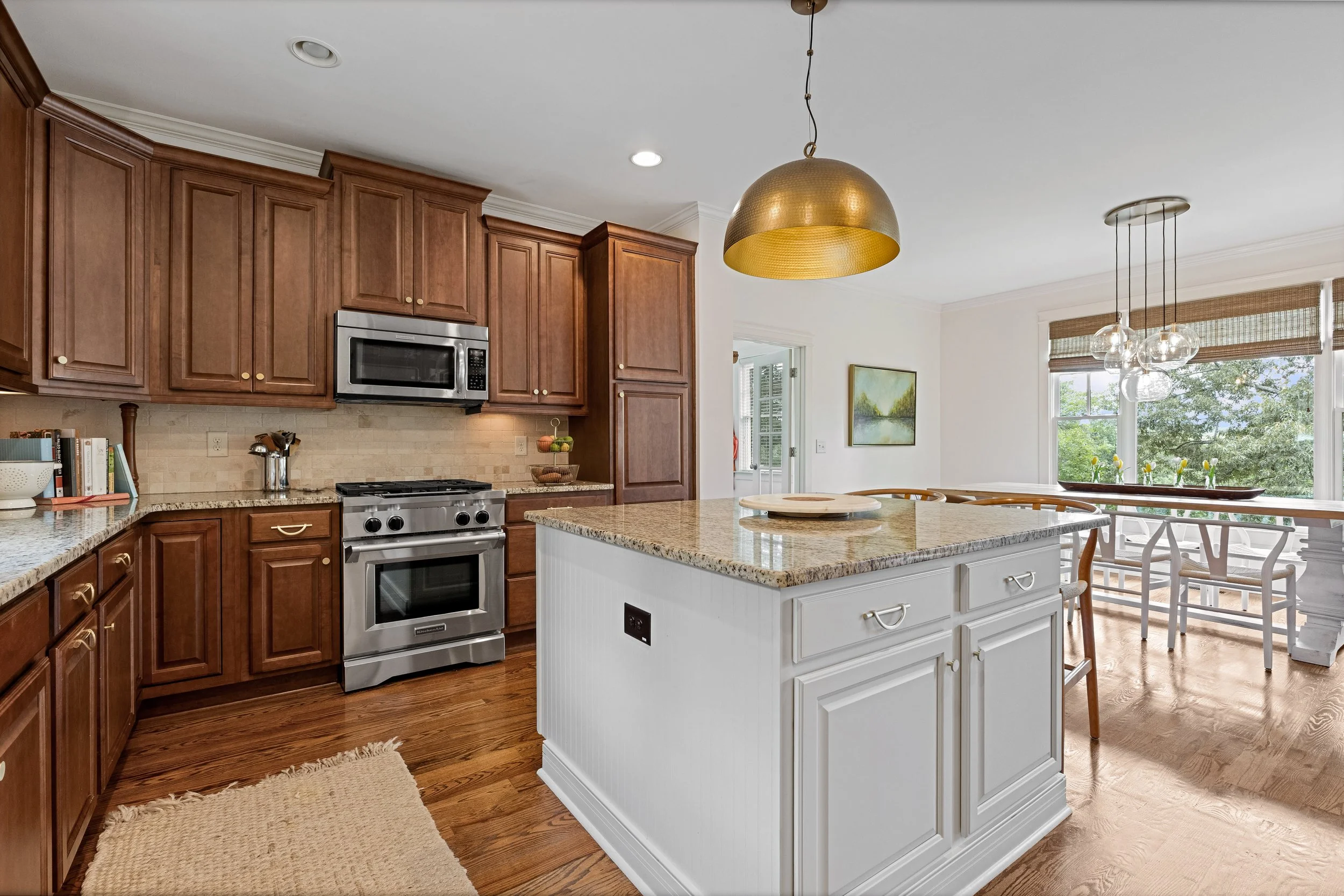 Bright kitchen with wooden cabinets, granite countertops, stainless steel oven and microwave, white island with drawers, hardwood floors, large window with shades, and modern light fixtures.