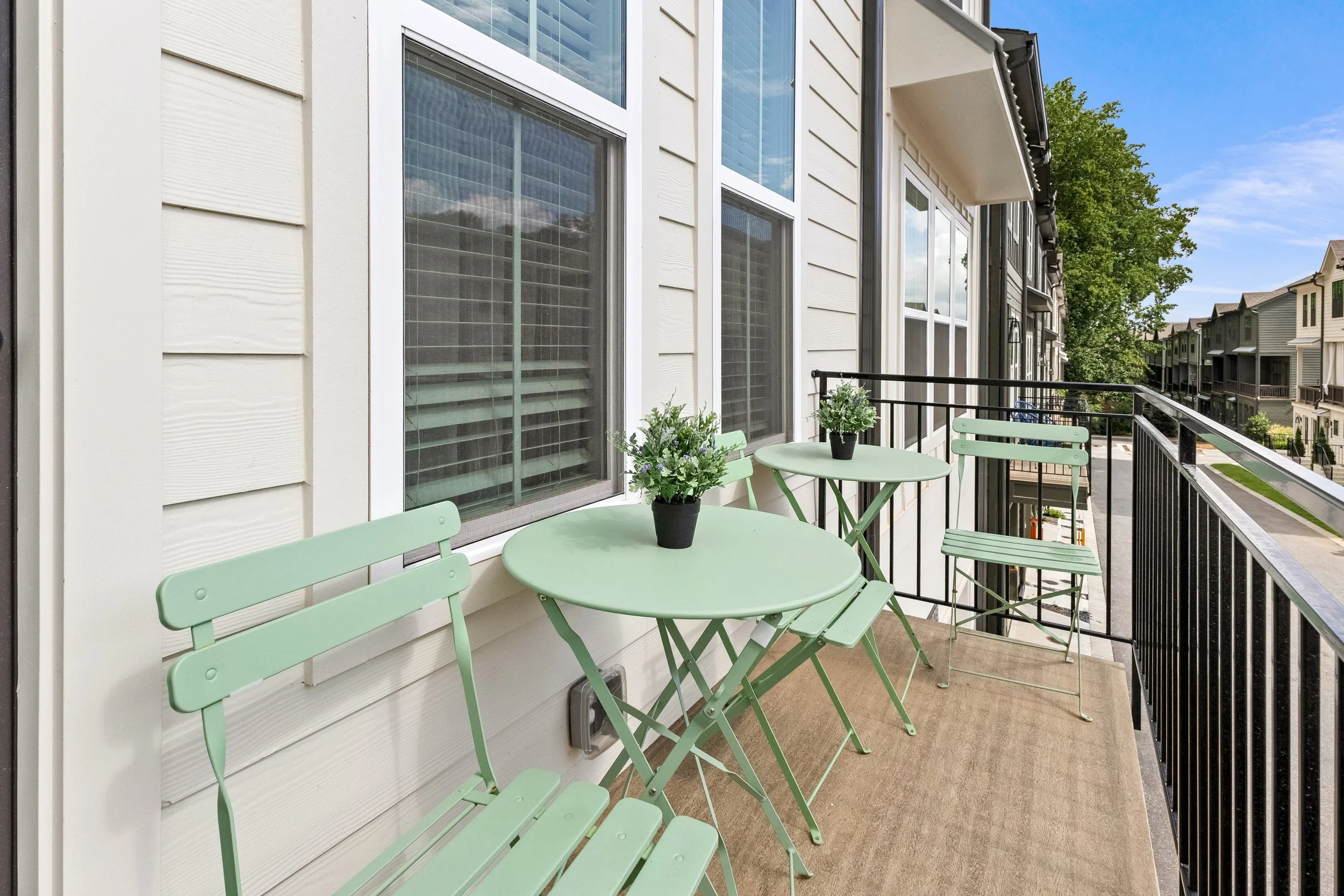 View of a balcony with light green metal chairs and a matching table with potted plants, overlooking buildings with a tree and blue sky in the background.