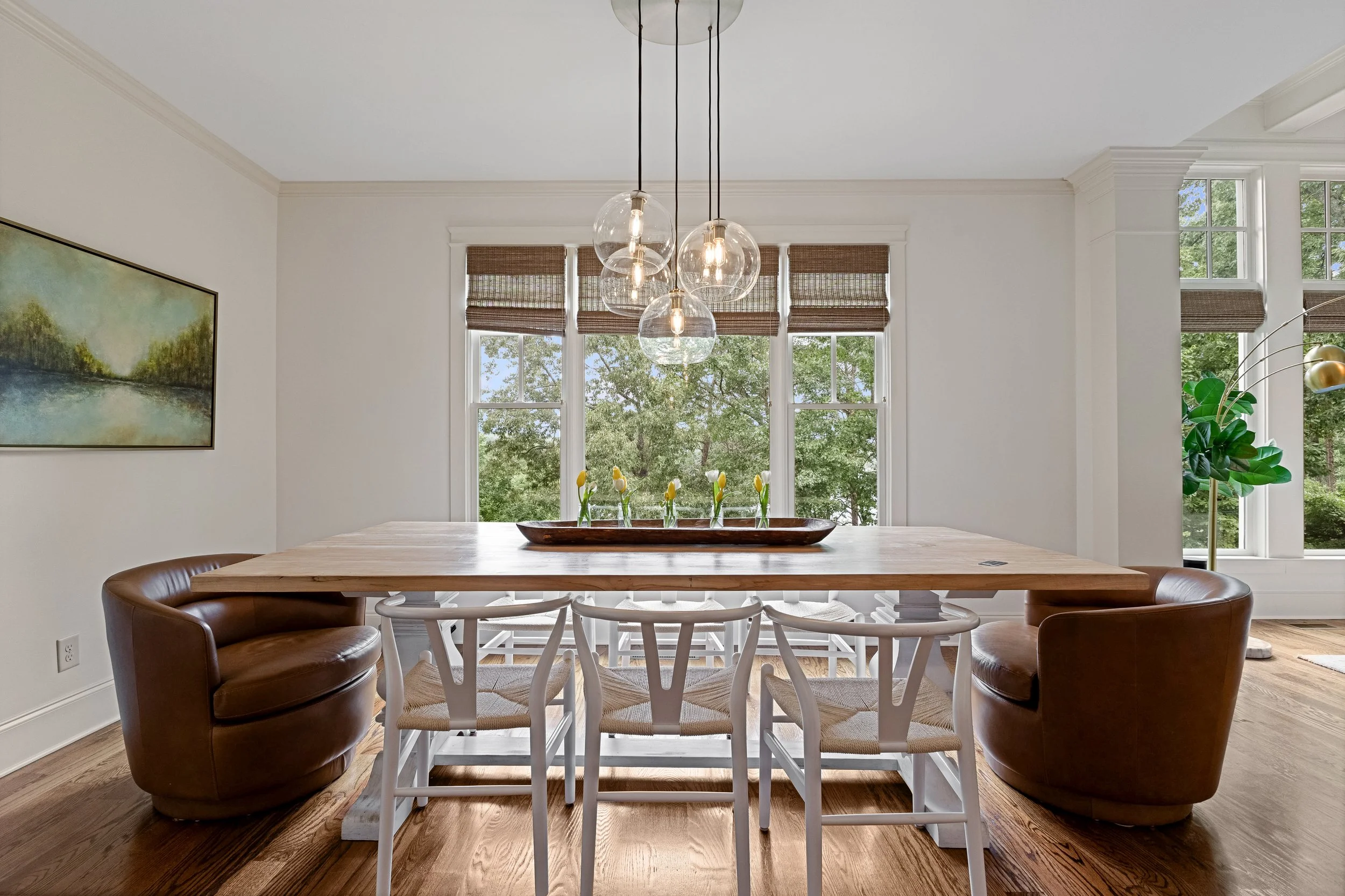Modern dining room with a wooden table, white chairs, brown leather armchairs, large windows with woven shades, and a pendant light fixture with five glass globes.