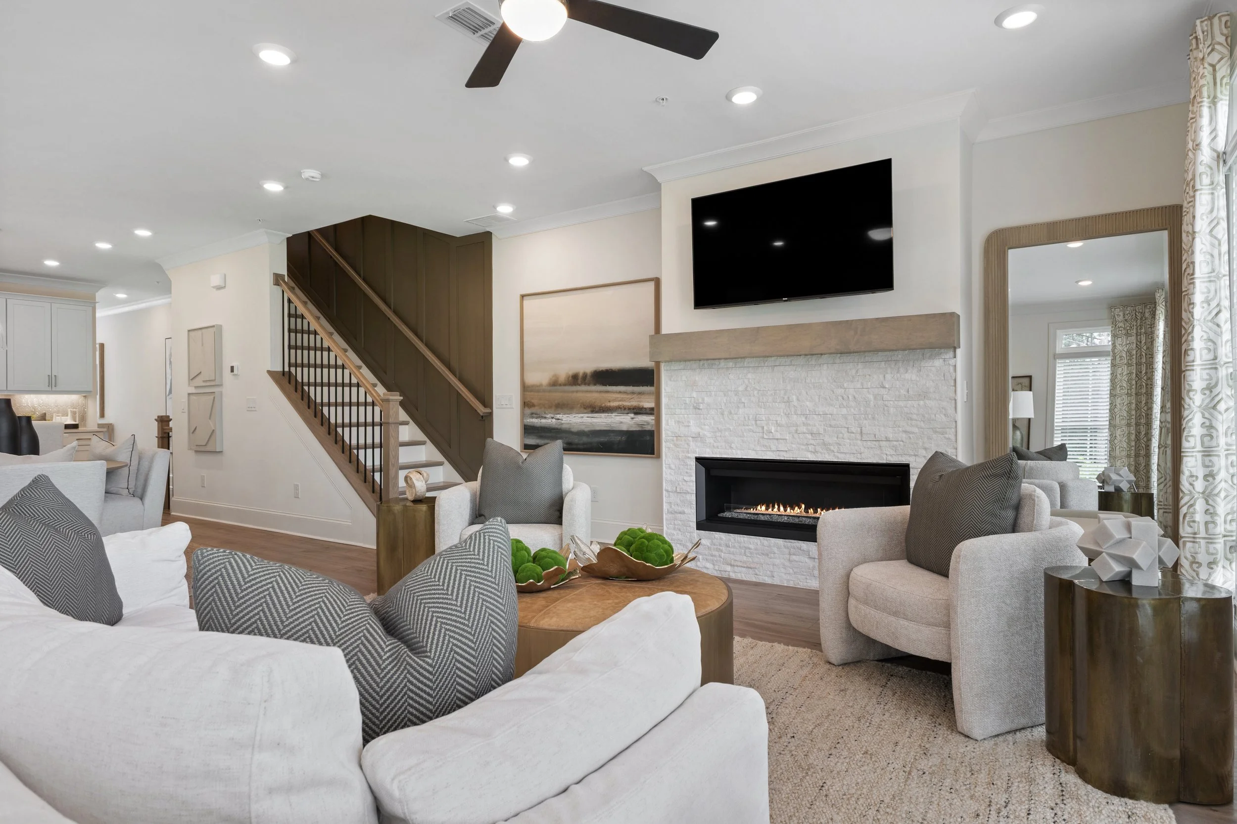 A modern living room with a white brick fireplace, mounted flat-screen TV, and a ceiling fan. Neutral-colored furniture including a white sofa and gray chairs, a wooden coffee table with green decorative objects, and large windows with patterned curtains.