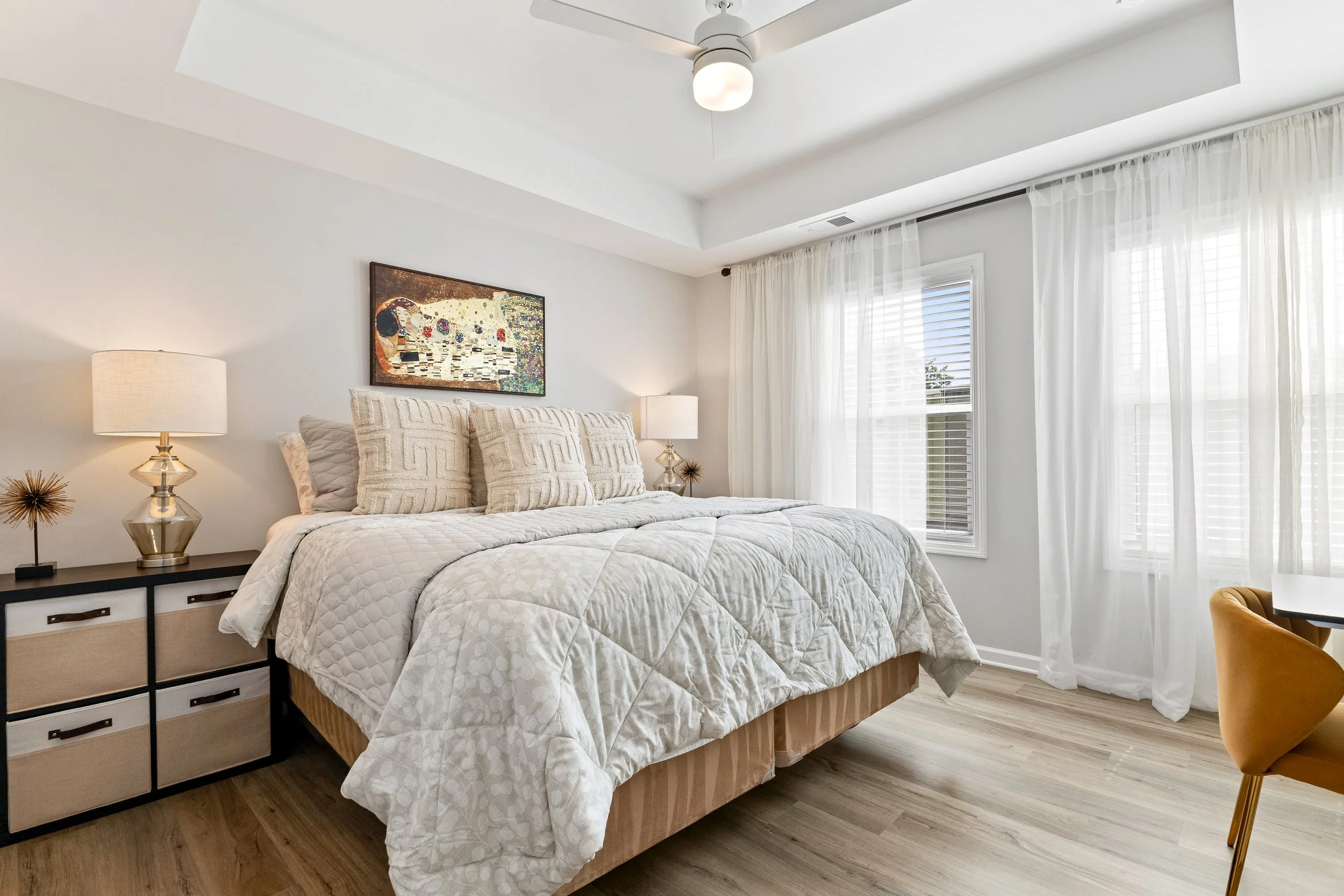 A modern bedroom with a large bed dressed in a textured beige comforter and matching pillows. There are two bedside lamps with brass bases, a black and beige dresser, and sheer white curtains over a window that allows natural sunlight. The room features light wood flooring and a framed colorful abstract art piece on the wall.