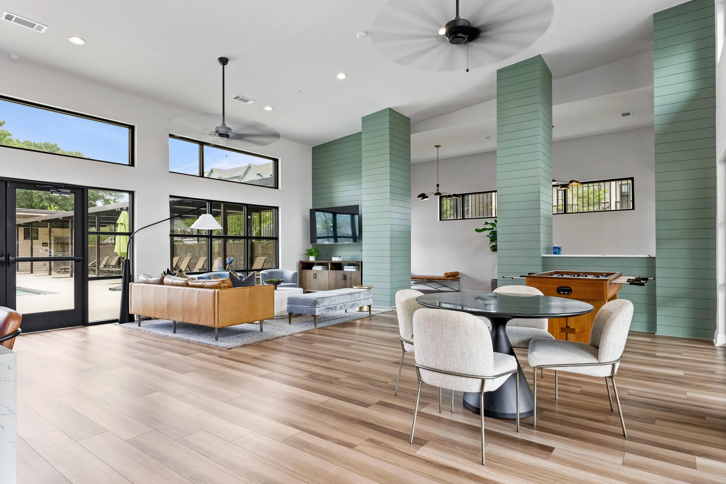 Bright, modern living room with high ceilings, large windows, and light wood flooring. Features include a tan sofa, a black round dining table with four beige upholstered chairs, green paneled columns, and a television on the wall. There are ceiling fans and sliding glass doors leading outside to a patio with outdoor furniture.