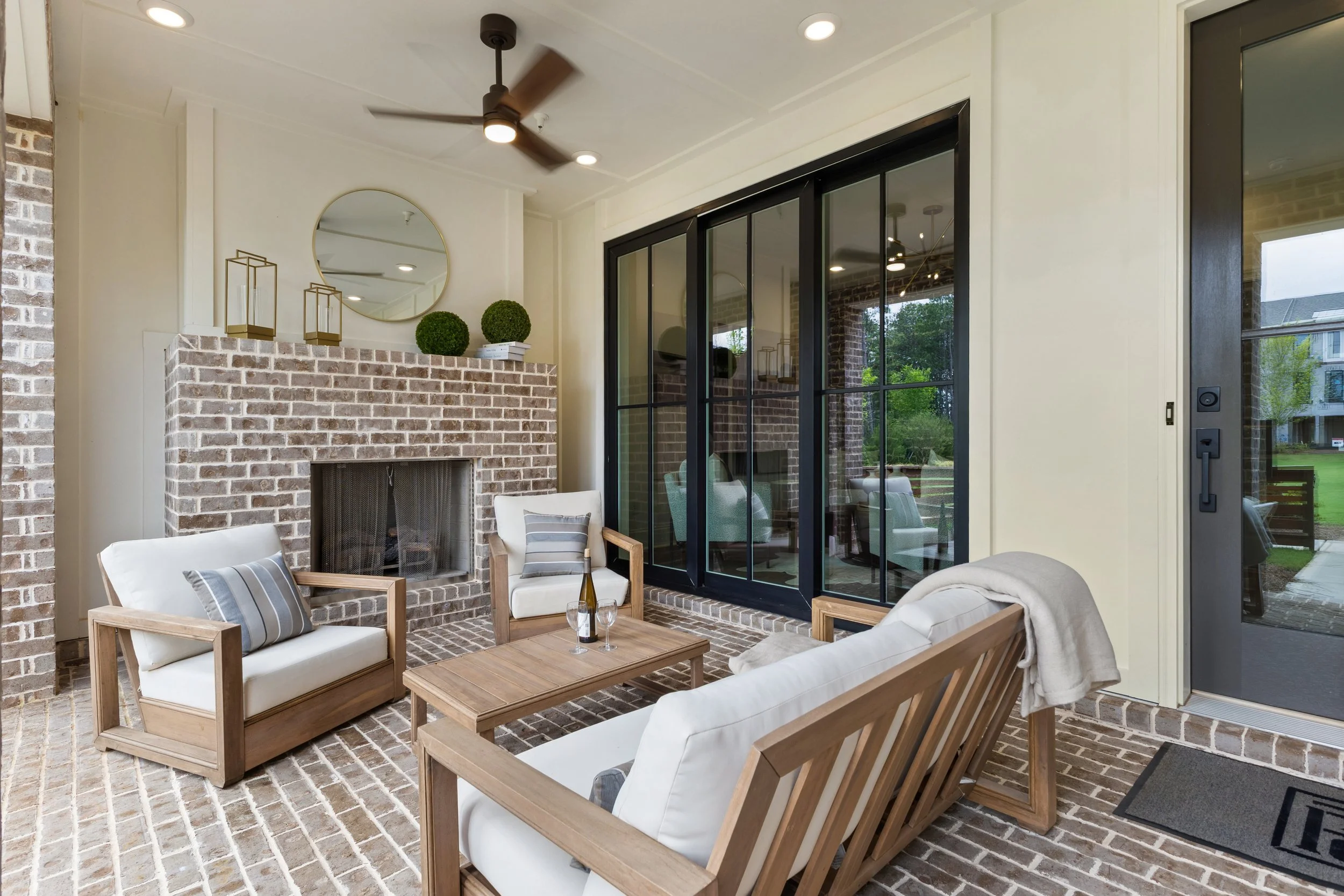 A cozy outdoor patio area with white cushioned wooden chairs, a brick fireplace, and a glass sliding door leading inside. Decor includes round green topiary plants, a mirror, and lanterns. The space has a brick and neutral-colored design.