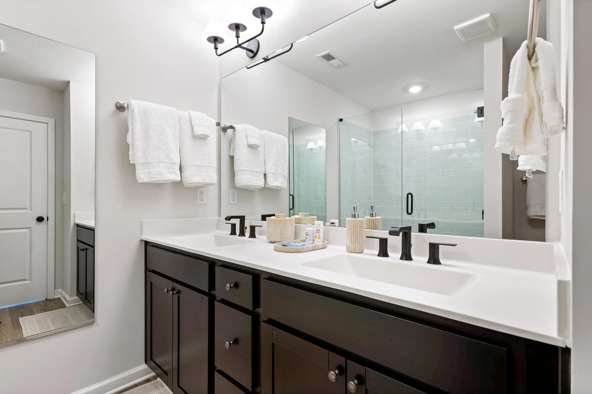 Modern bathroom with a double vanity, large mirror, black fixtures, white countertop, and towels hanging on towel bars. There is a glass shower enclosure with white subway tiles in the background.