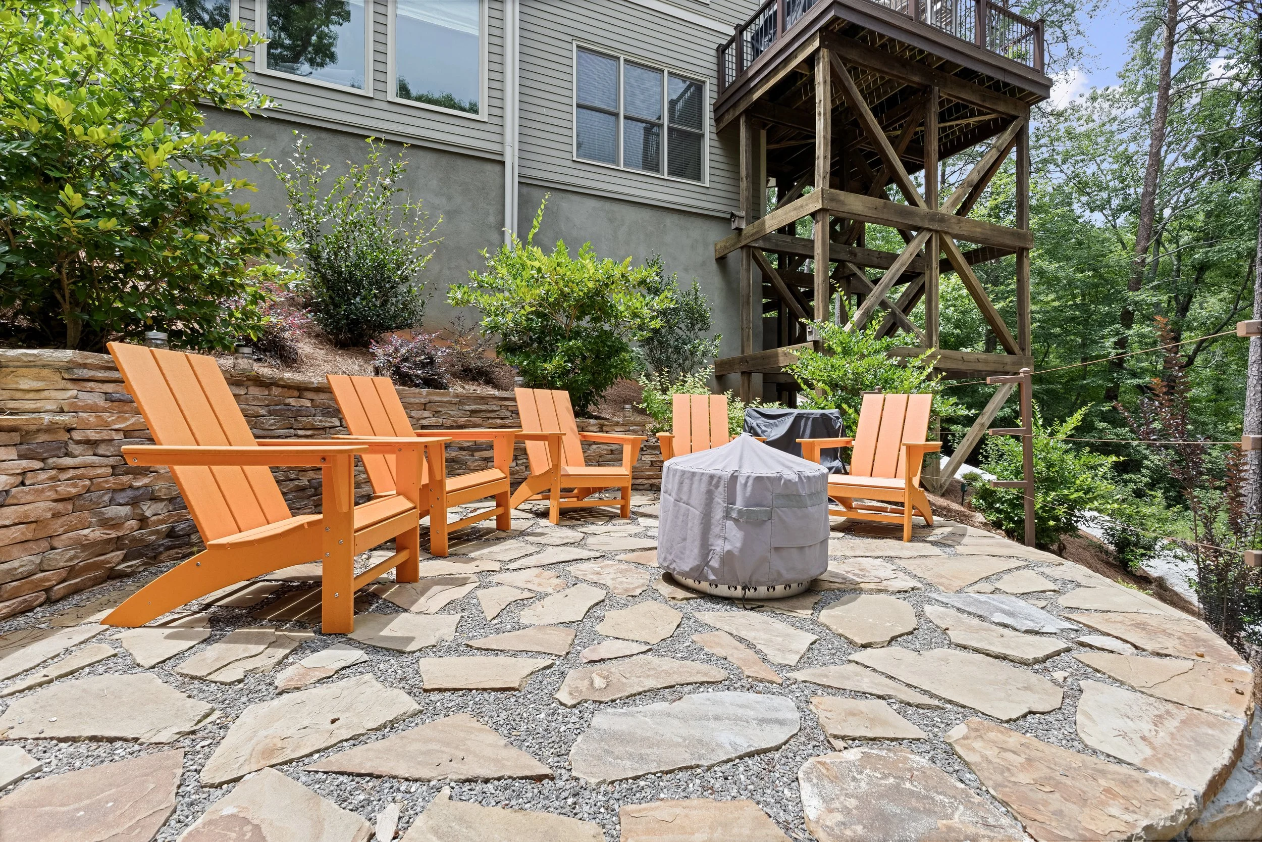 Backyard patio area with Adirondack chairs, a grill covered with a tarp, stone paving, and a wooden deck above surrounded by trees and bushes.
