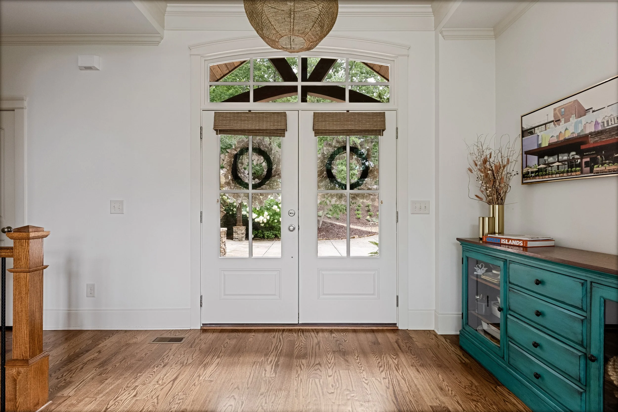 Entryway with white door and window panels, view of greenery and trees outside, wicker shades on door, a blue cabinet with books and decorative items, dried plants in a gold vase, and wall art.