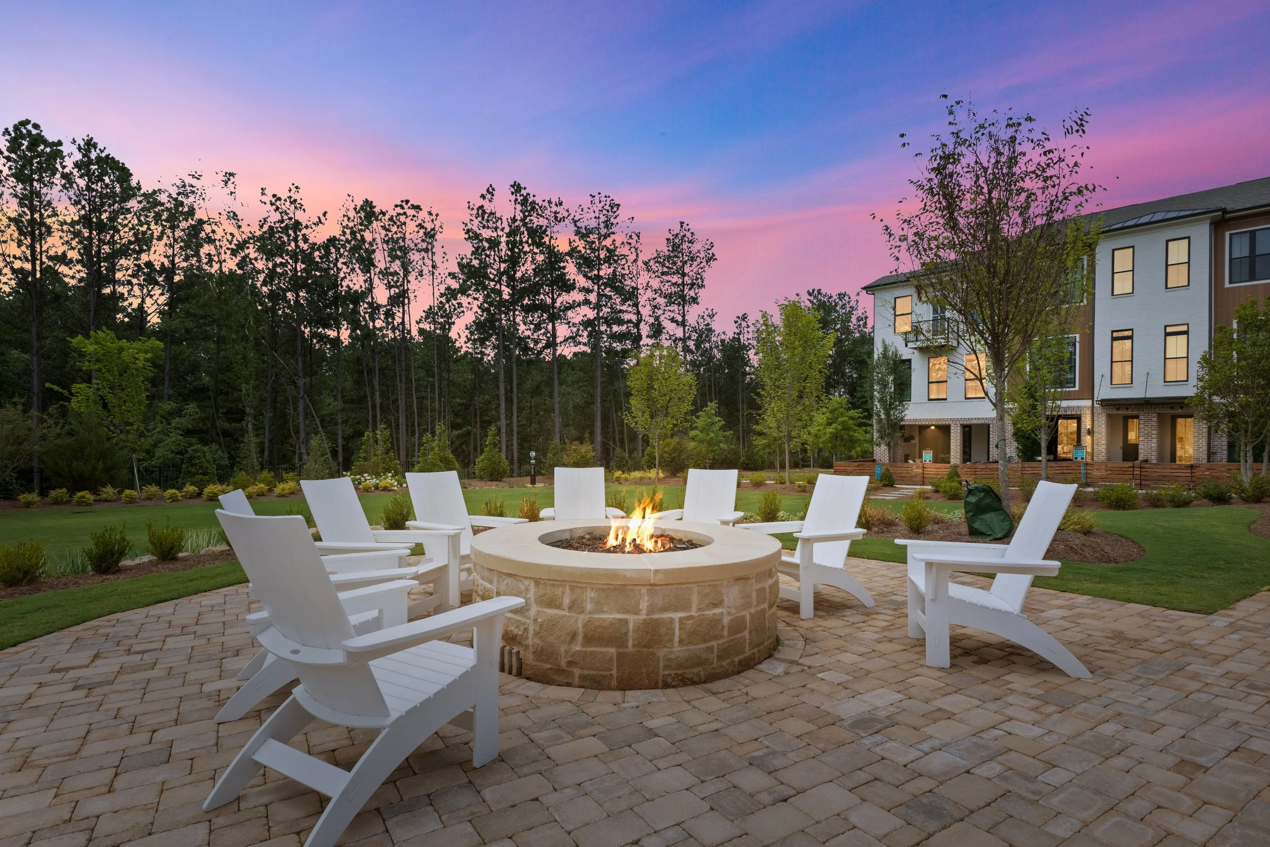 An outdoor patio with a fire pit surrounded by white chairs. In the background, there are trees, a grassy area, and a residential building, with a colorful sunset sky overhead.