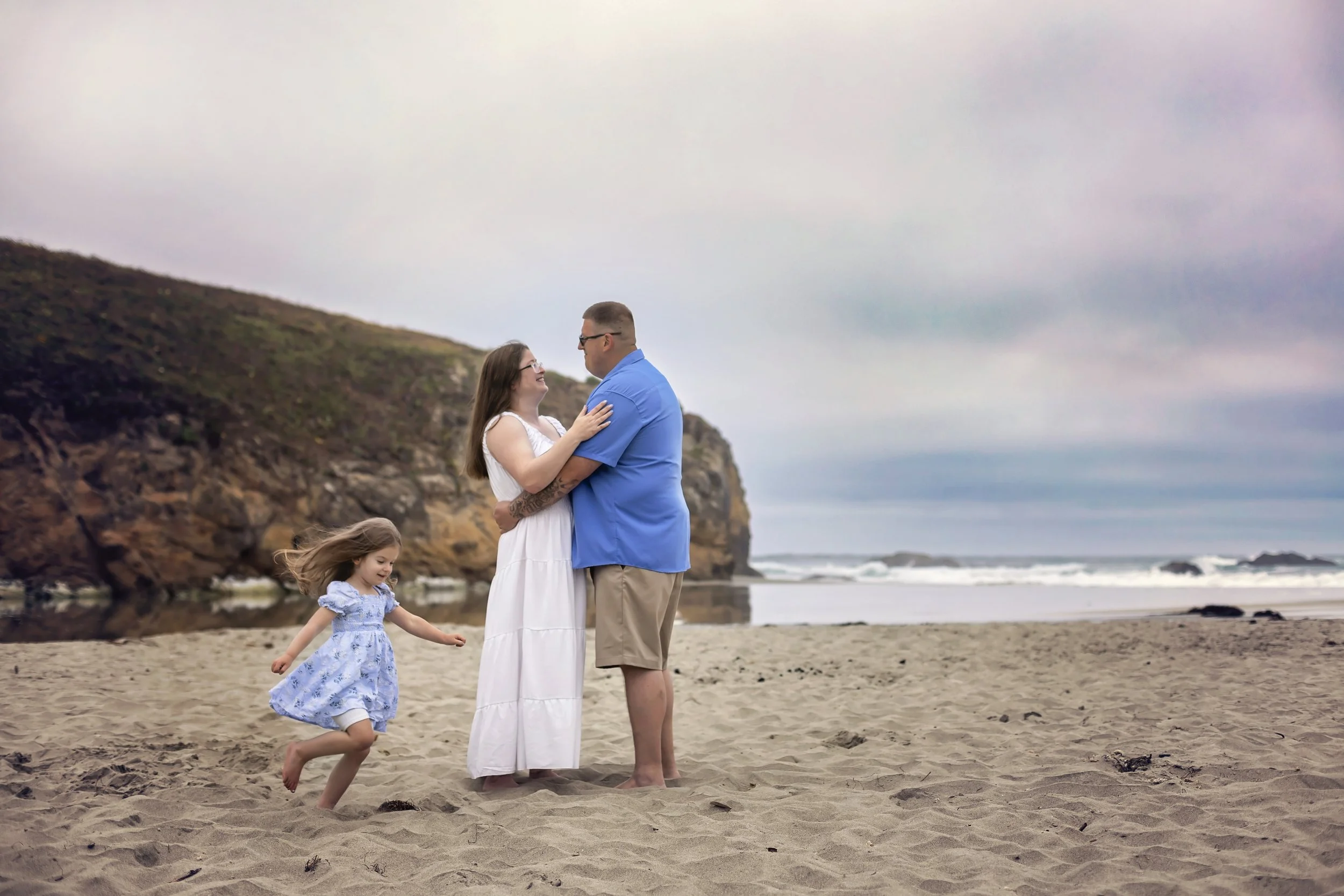 A family of four having fun on the beach. A woman in a white dress, a man in a blue shirt, and two young girls in blue dresses are present. The woman and man are looking at each other while the girls are playing and running on the sand. Cliffs and the ocean can be seen in the background on a cloudy day.