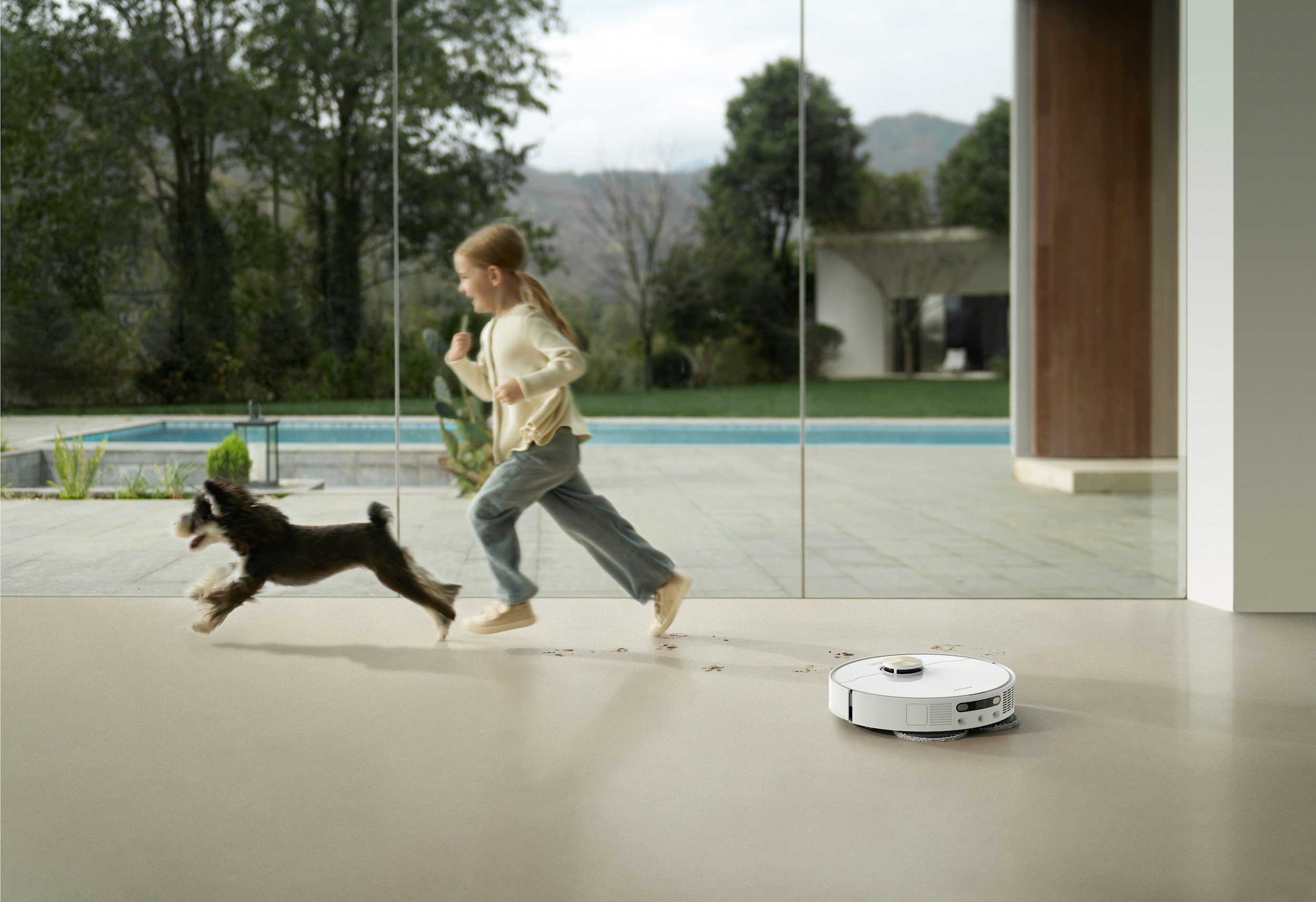 A young girl running with a dog near a glass wall with an ocean and green trees outside.