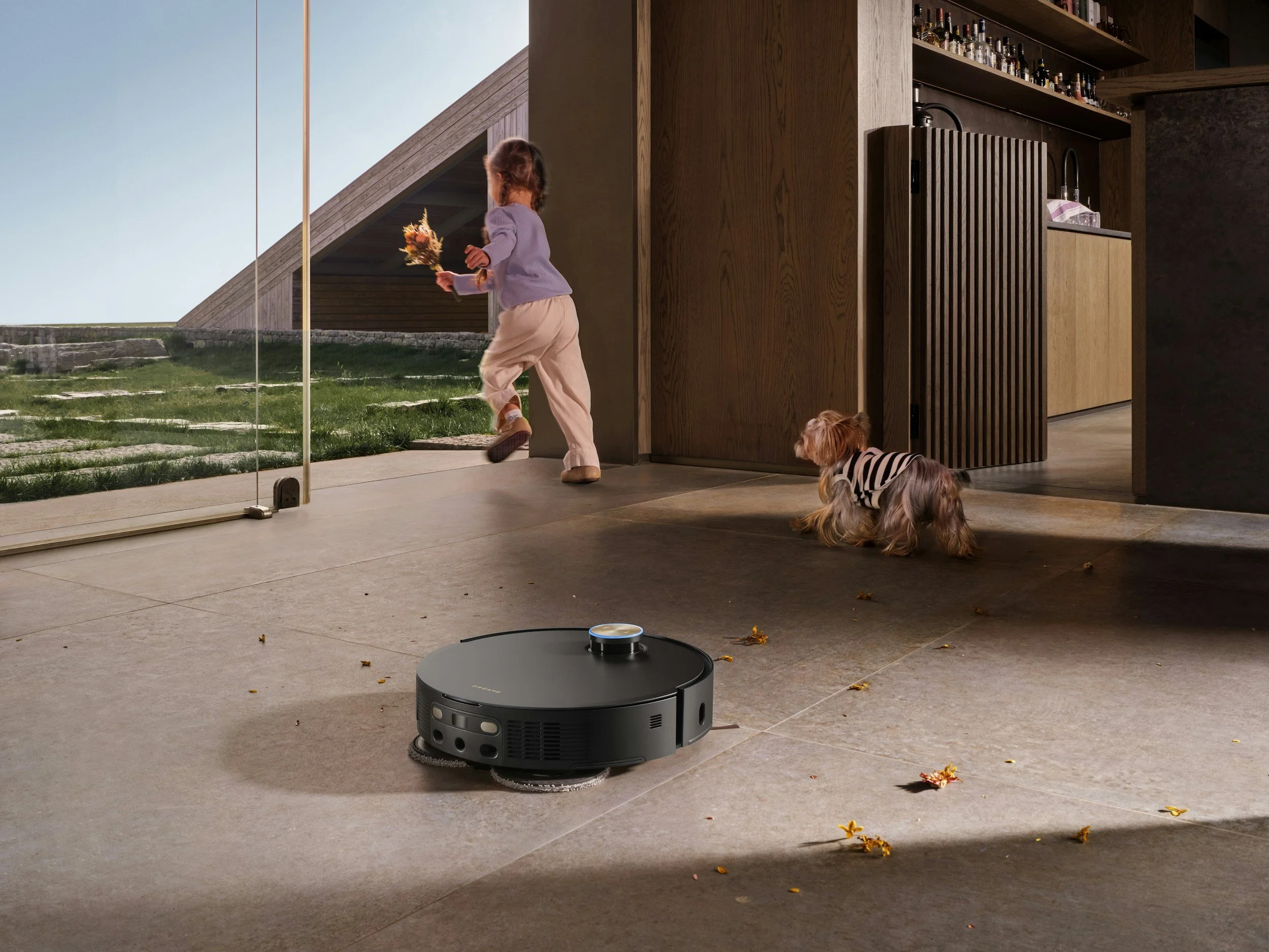 A young girl with her hair in a braid, wearing a lavender sweater and beige pants, holding a bouquet of flowers and walking near a glass door, with two small dogs on the floor inside the house. The interior features wooden cabinetry and a black robot vacuum cleaner on the beige tiled floor, with some leaves and flowers scattered.