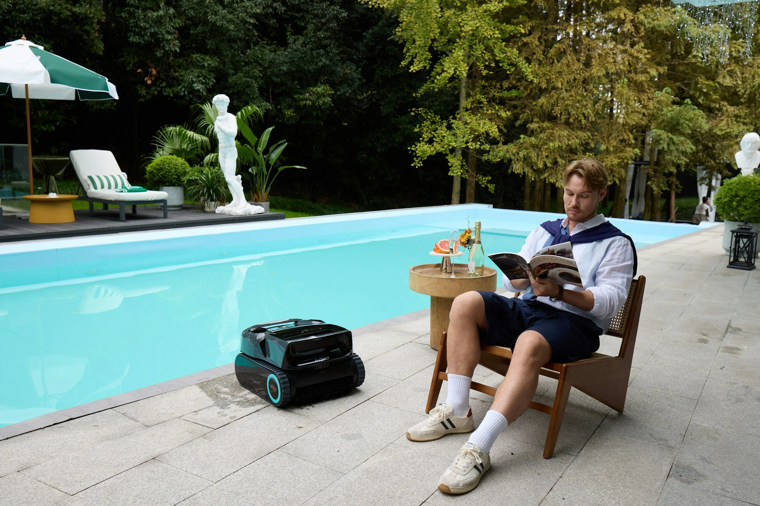 A man sitting on a wooden chair by a swimming pool, reading a magazine. He is wearing a white shirt with a navy blue sweater draped over his shoulders, shorts, white socks, and sneakers. There is a drink with grapefruit slices and a bottle of champagne on a small table beside him. A robotic vacuum cleaner is on the ground nearby. The background includes green trees, decorative statues, a lounge chair with striped cushion, a yellow side table, and an umbrella.